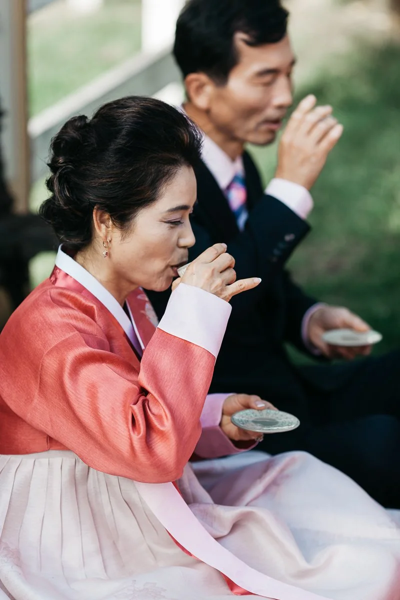 A woman and a man, dressed in traditional Korean hanbok attire, sitting outdoors eating from small dishes.