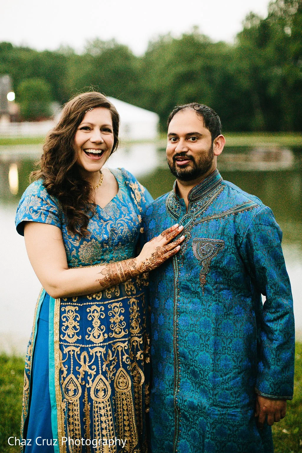 A couple dressed in traditional Indian clothing standing outdoors by a lake with trees in the background. The woman is smiling, wearing a blue and gold embroidered dress with mehndi on her hands. The man has facial hair, wearing a blue embroidered kurta.