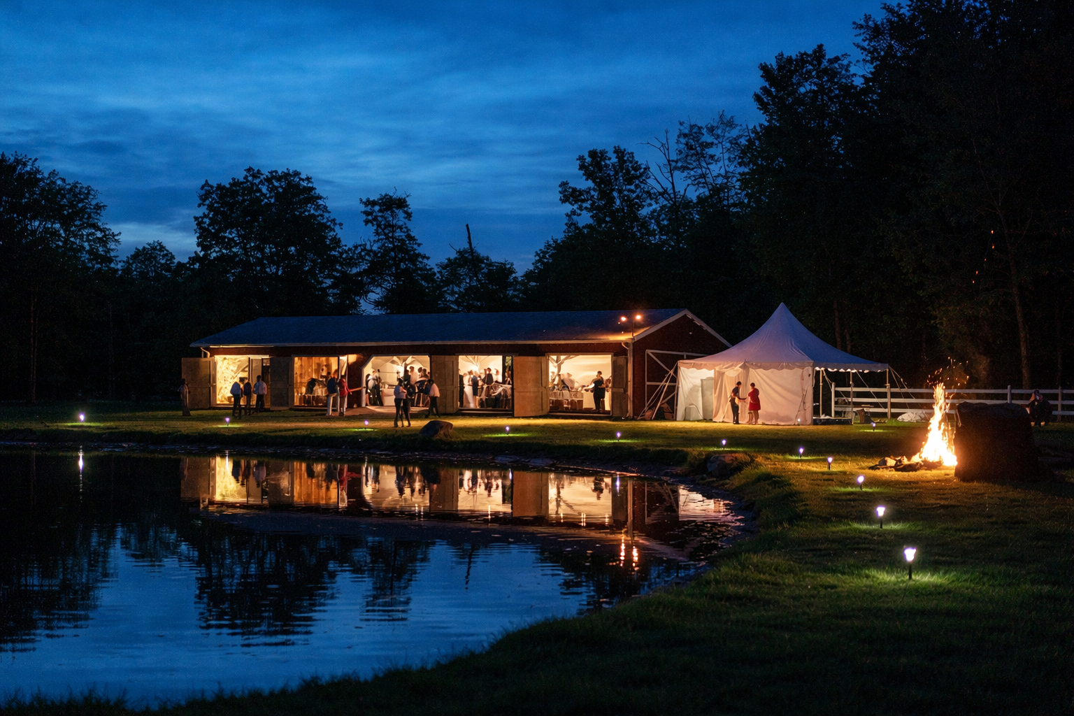 Night scene of a social event with people inside a well-lit barn and outside a white tent, near a pond with reflections, surrounded by trees and a fire on the ground.