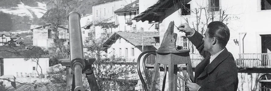 A man painting a Santa hat on a witch's hat on a chair outdoors with buildings in the background.
