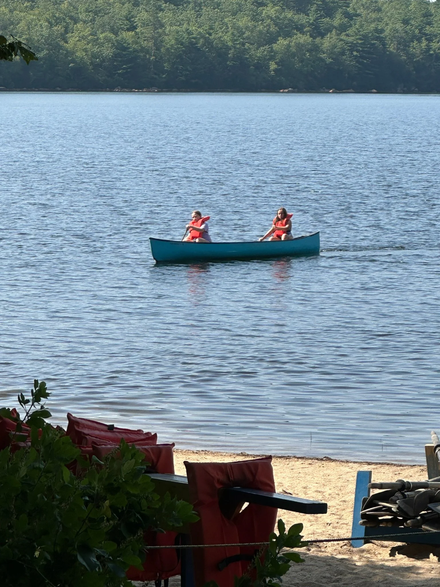 Two people in a blue canoe on Yawgoog Pond wearing red life jackets on a lake, with trees in the background and chairs on the shore.