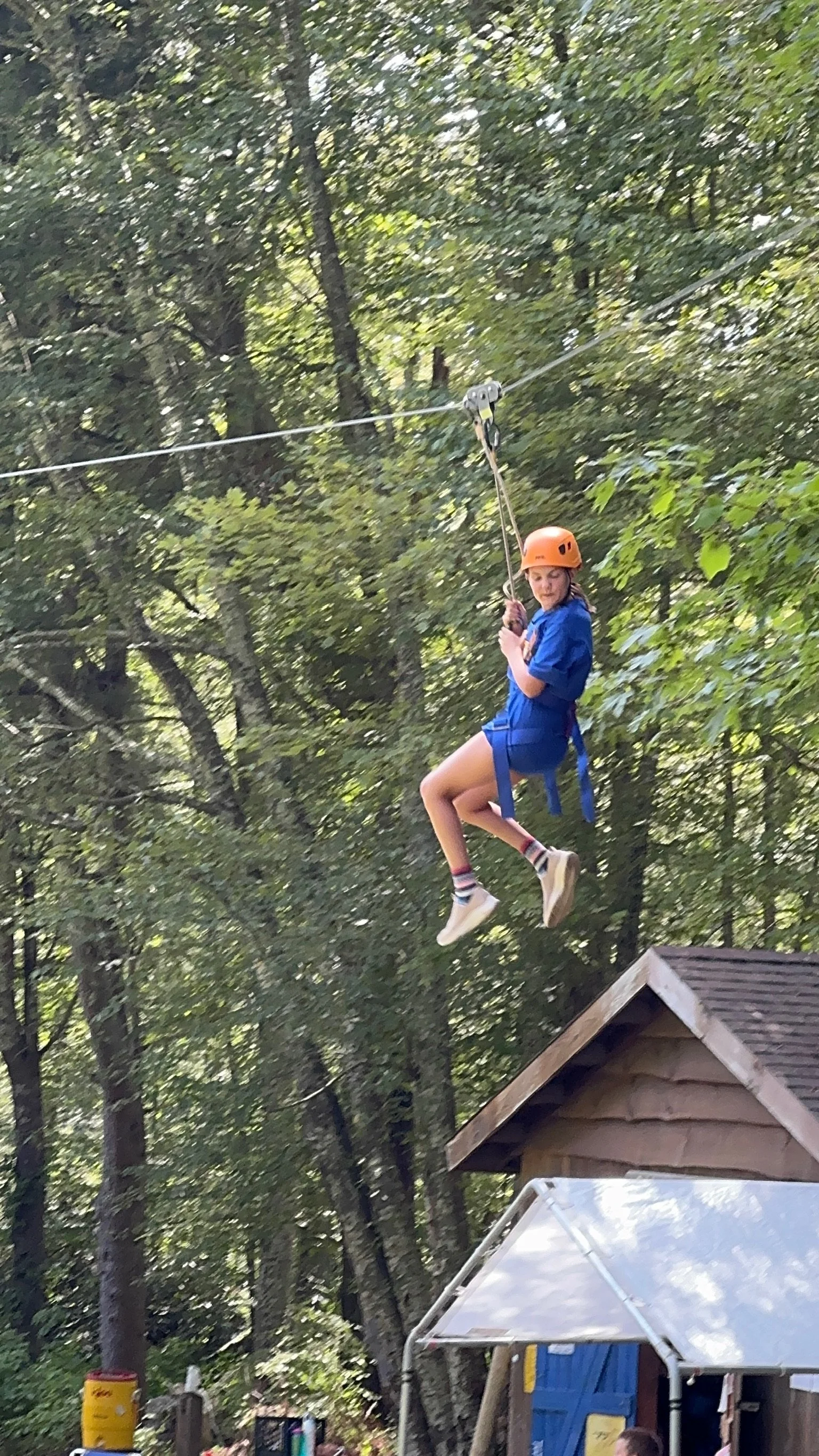 Girl in a blue outfit and orange helmet riding a zip line in a wooded outdoor area.