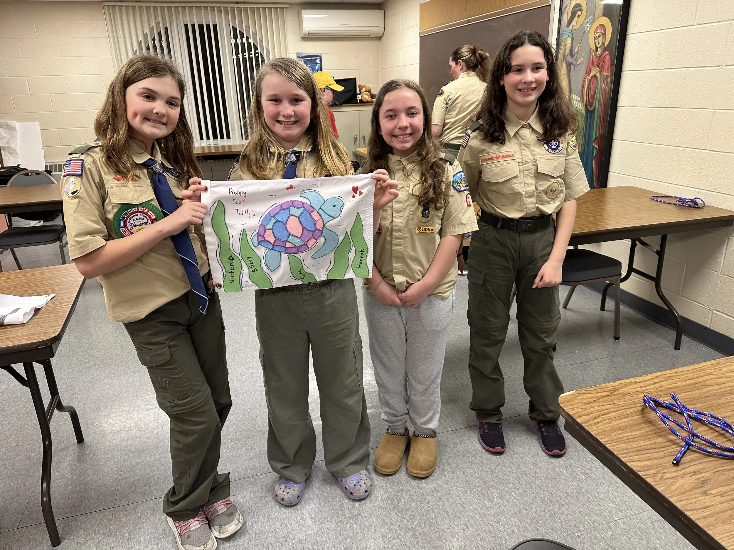 Four Girl Scouts in uniform standing in a room, holding a colorful drawing of a turtle with the words 'Preppy  Turtle's'. Other Girl Scouts and a leader are visible in the background, with tables and chairs around.