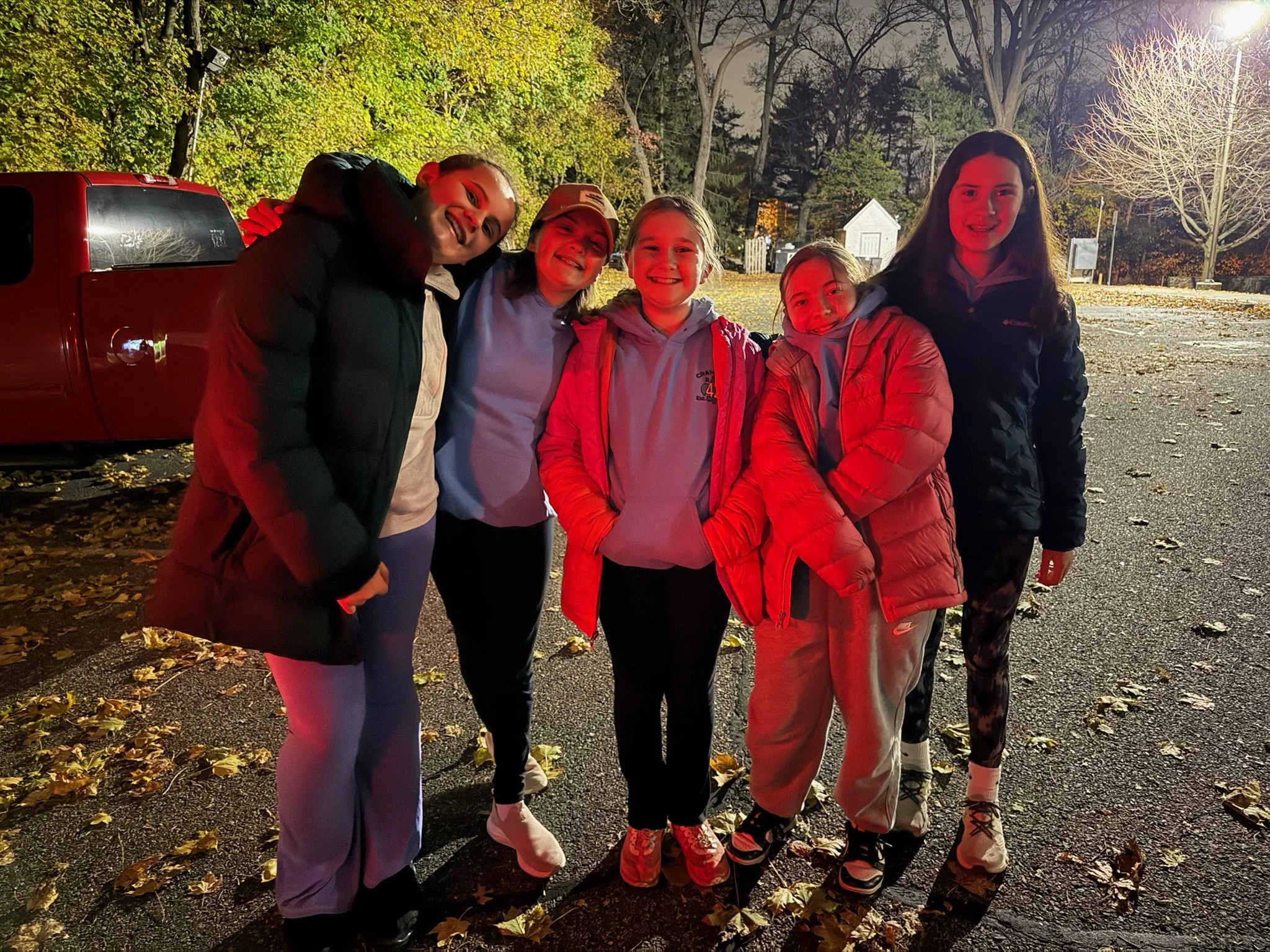 Five young women standing together outdoors at night, smiling, dressed in jackets and athletic wear, with a red truck and trees in the background.