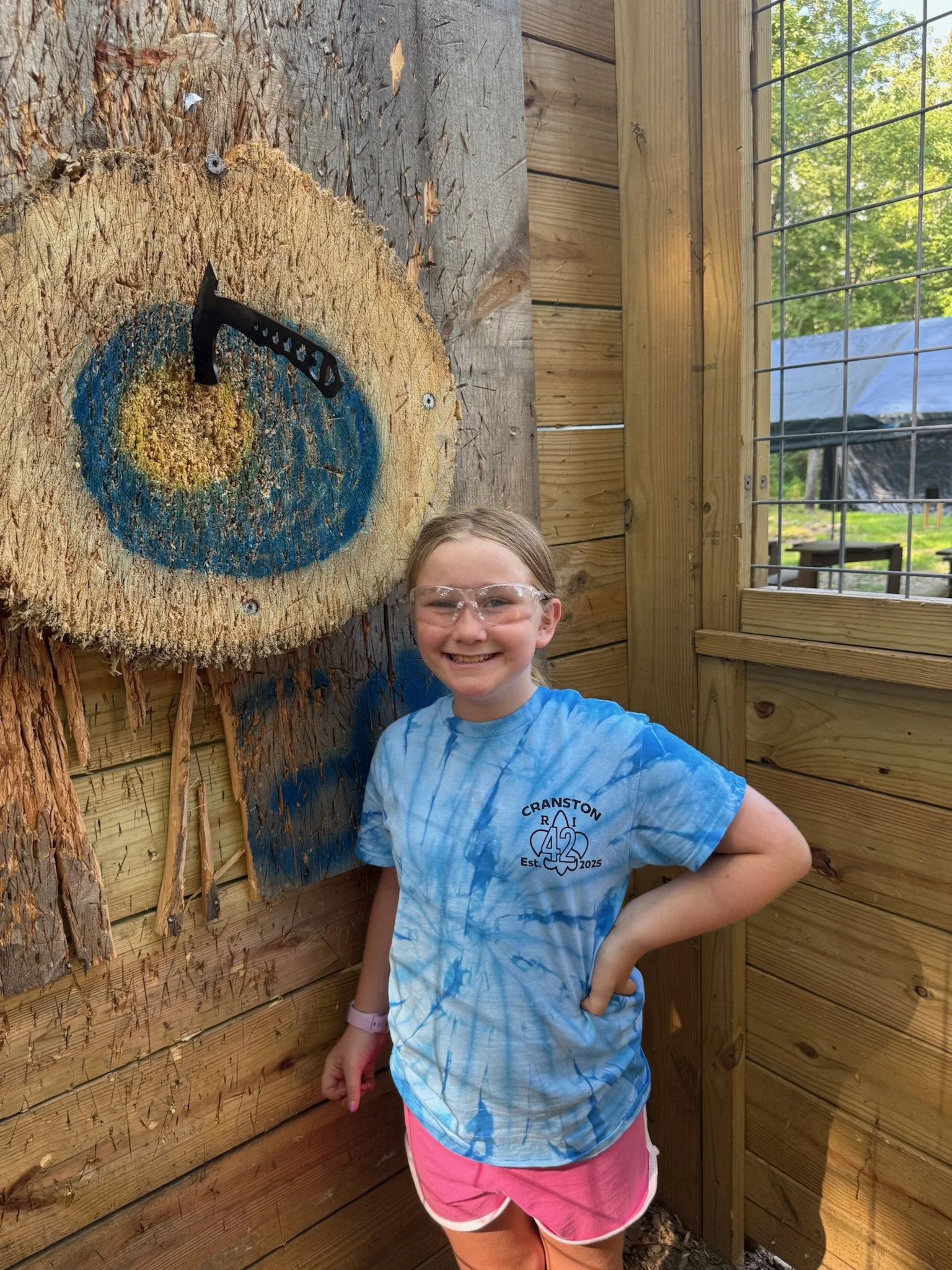 A young girl smiling and standing next to a large section of a tree stump with a blue and yellow painted target on it and a small axe embedded in the wood. The girl is wearing safety glasses, a blue tie-dye t-shirt, and pink shorts. She is in an outdoor enclosure with wooden walls and a wire mesh window, with trees in the background.