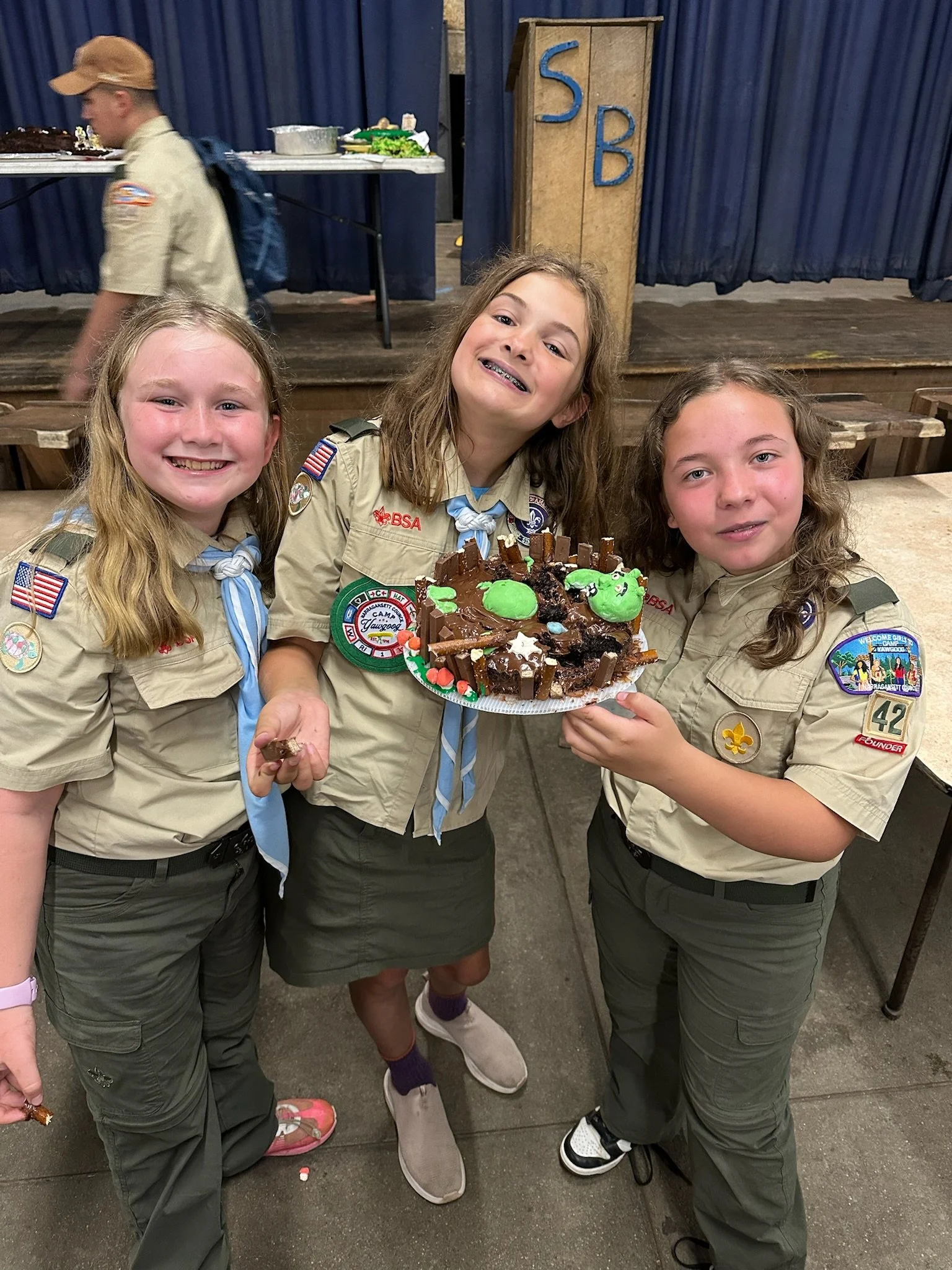 Three Girl Scouts in uniform holding a decorated cake outdoors in a hall with a stage and blue curtains.