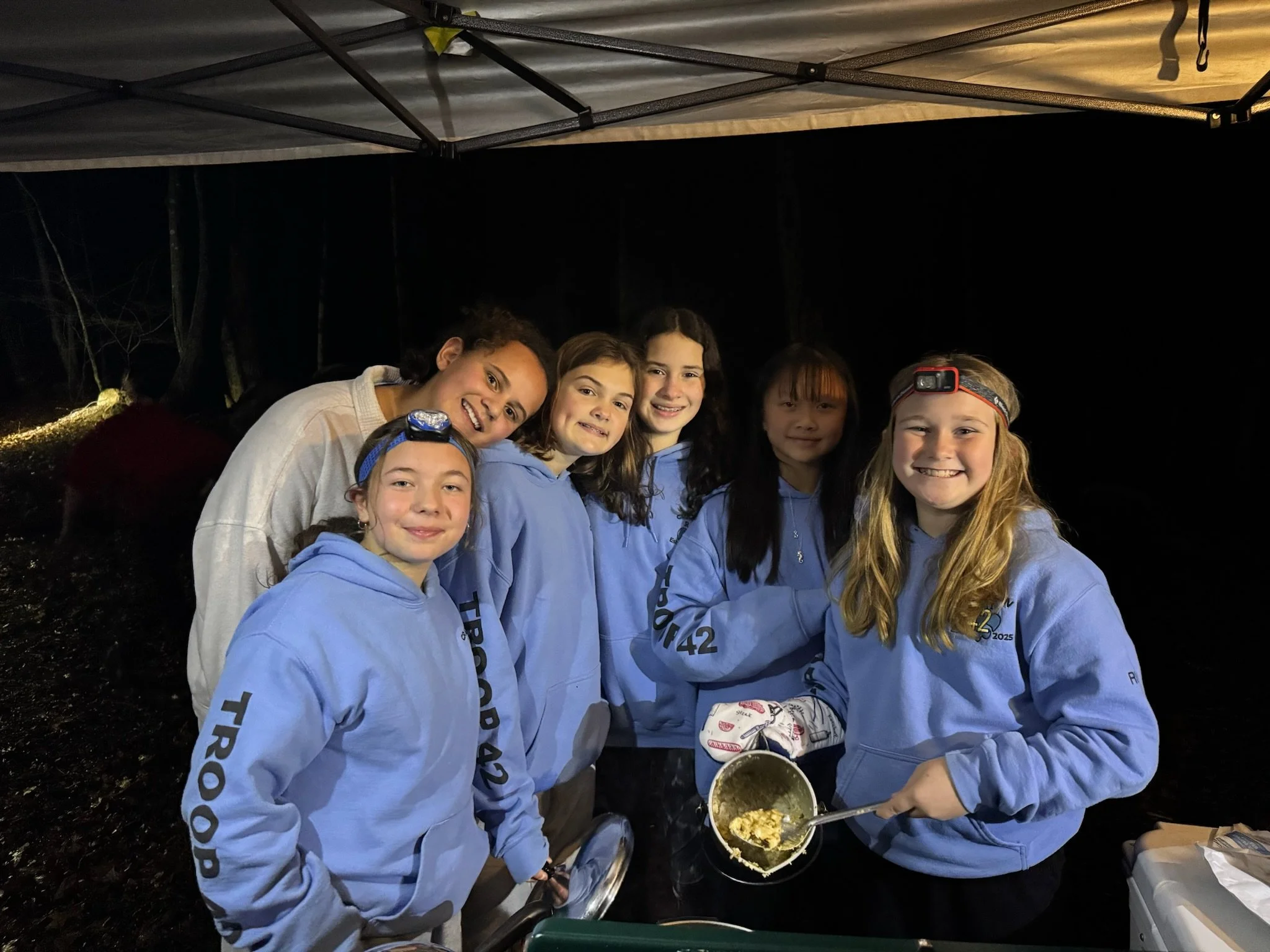 A group of six girls wearing matching blue hoodies, some with headlamps, standing under a canopy at night. One girl is holding a pot of cooked food, smiling at the camera.