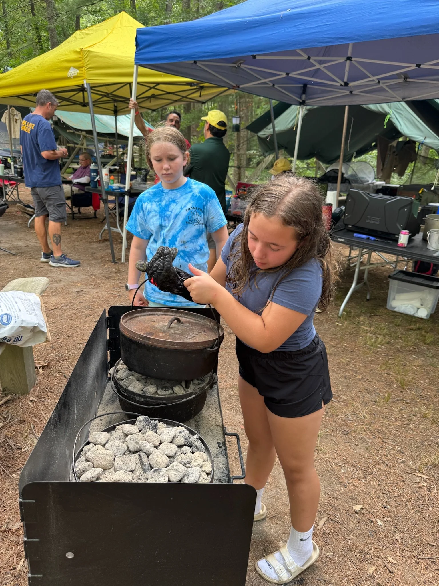A girl cooking over a campfire grill with rocks, surrounded by tents at a campsite. Two girls watch, with tents and trees in the background.