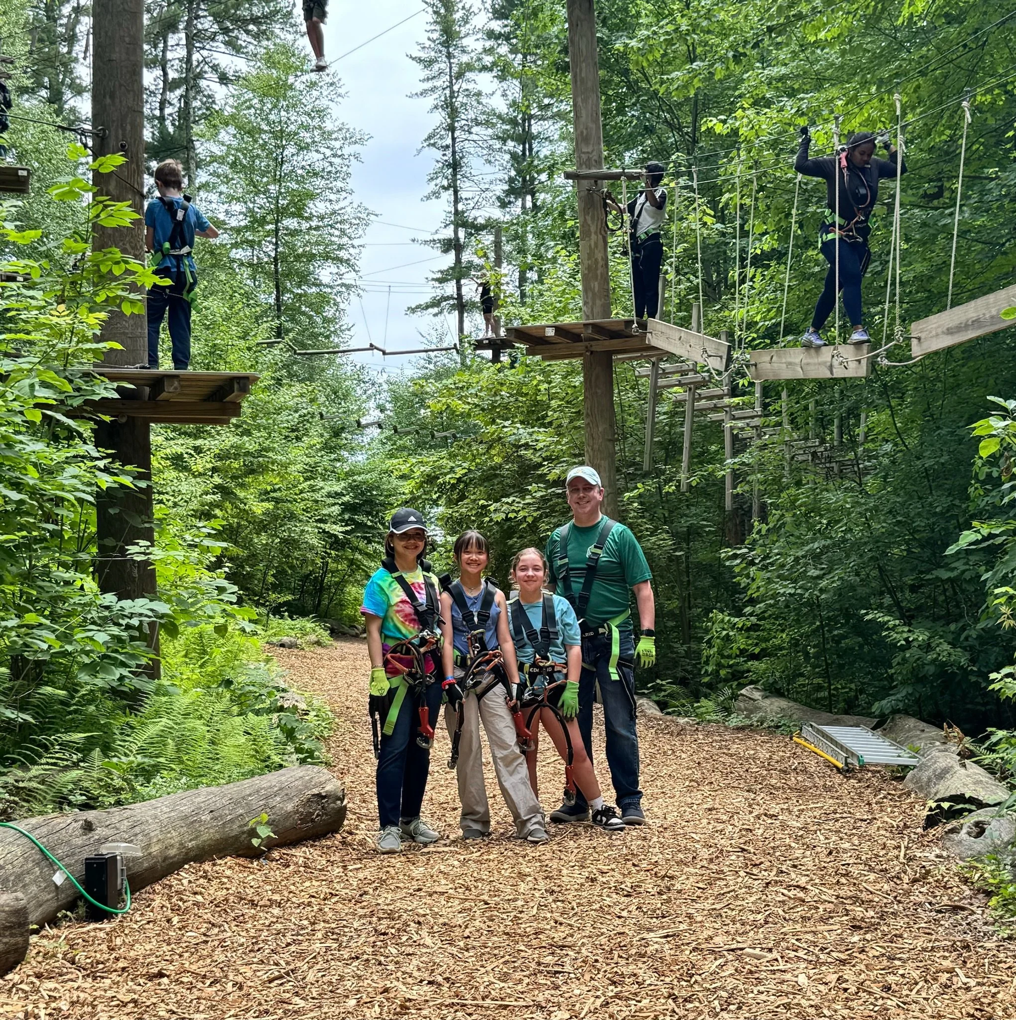 Group of four people standing on a trail in a forested outdoor adventure park with harnesses, surrounded by trees and outdoor activity ropes course.