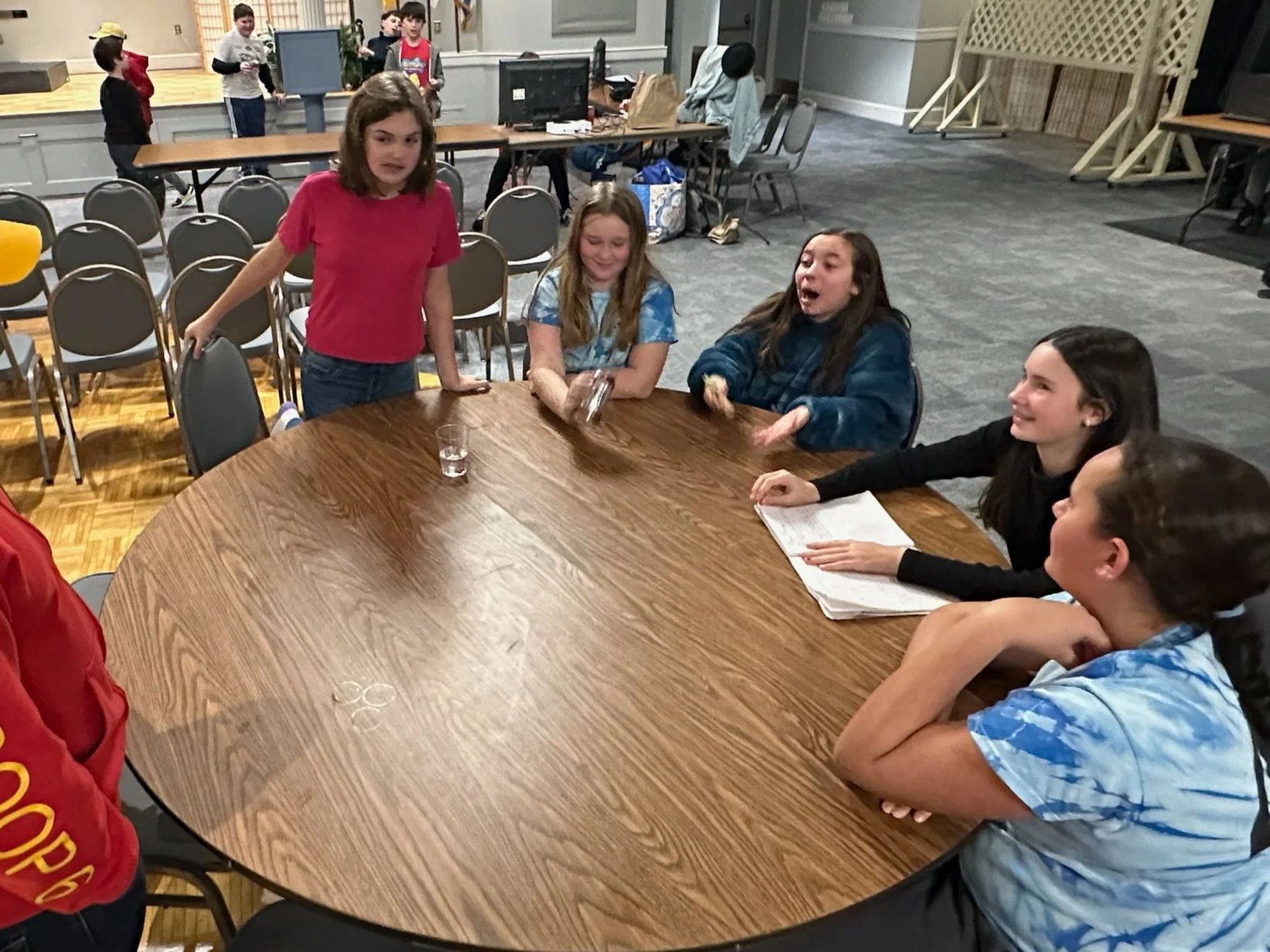 A group of children sitting and standing around a large round wooden table in a room with chairs, computers, and other children in the background.