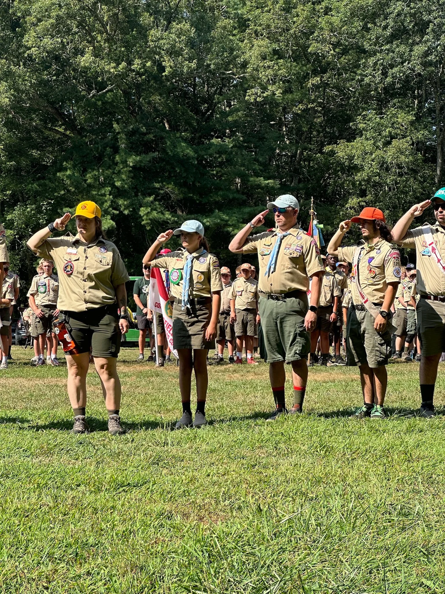 A group of Scouts BSA Scouts and Scout leaders standing on grass and saluting during the closing ceremony at Camp Yawgoog in Rockville RI, with trees in the background.