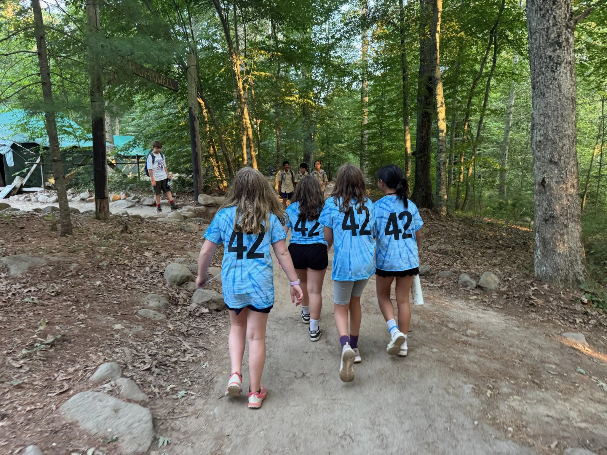 A group of young girls with matching blue sports jerseys, each with the number 42 on the back, walking on a dirt trail through a wooded forest area, with some rocks and trees surrounding the path.