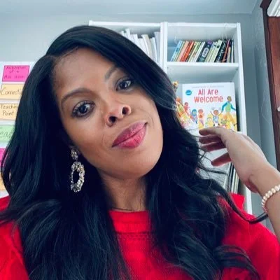 Woman with long black hair wearing a red top and earrings, in front of bookshelves with colorful books and decorations.