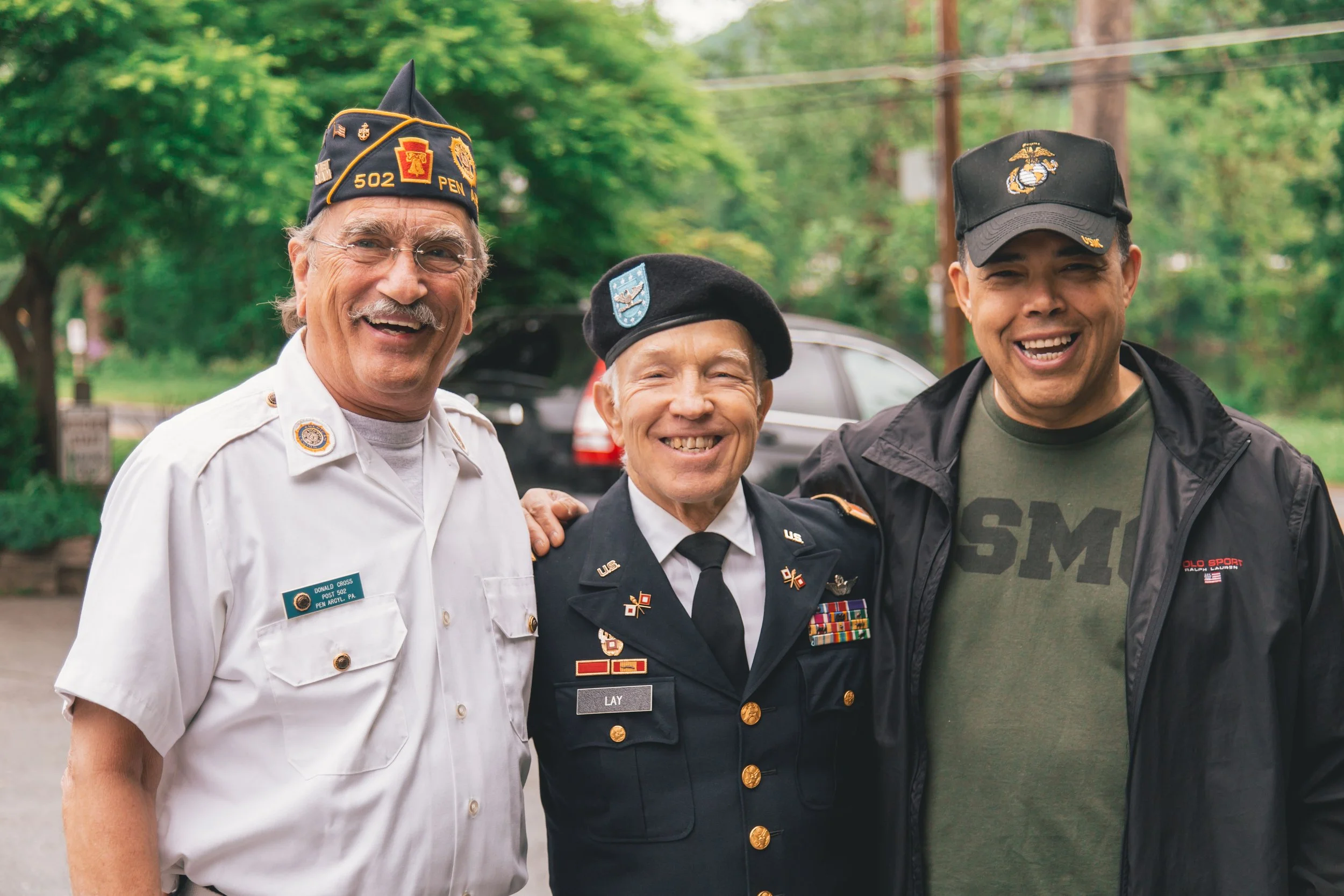 Three men smiling outdoors, wearing military uniforms and cap, with trees and a car in the background.