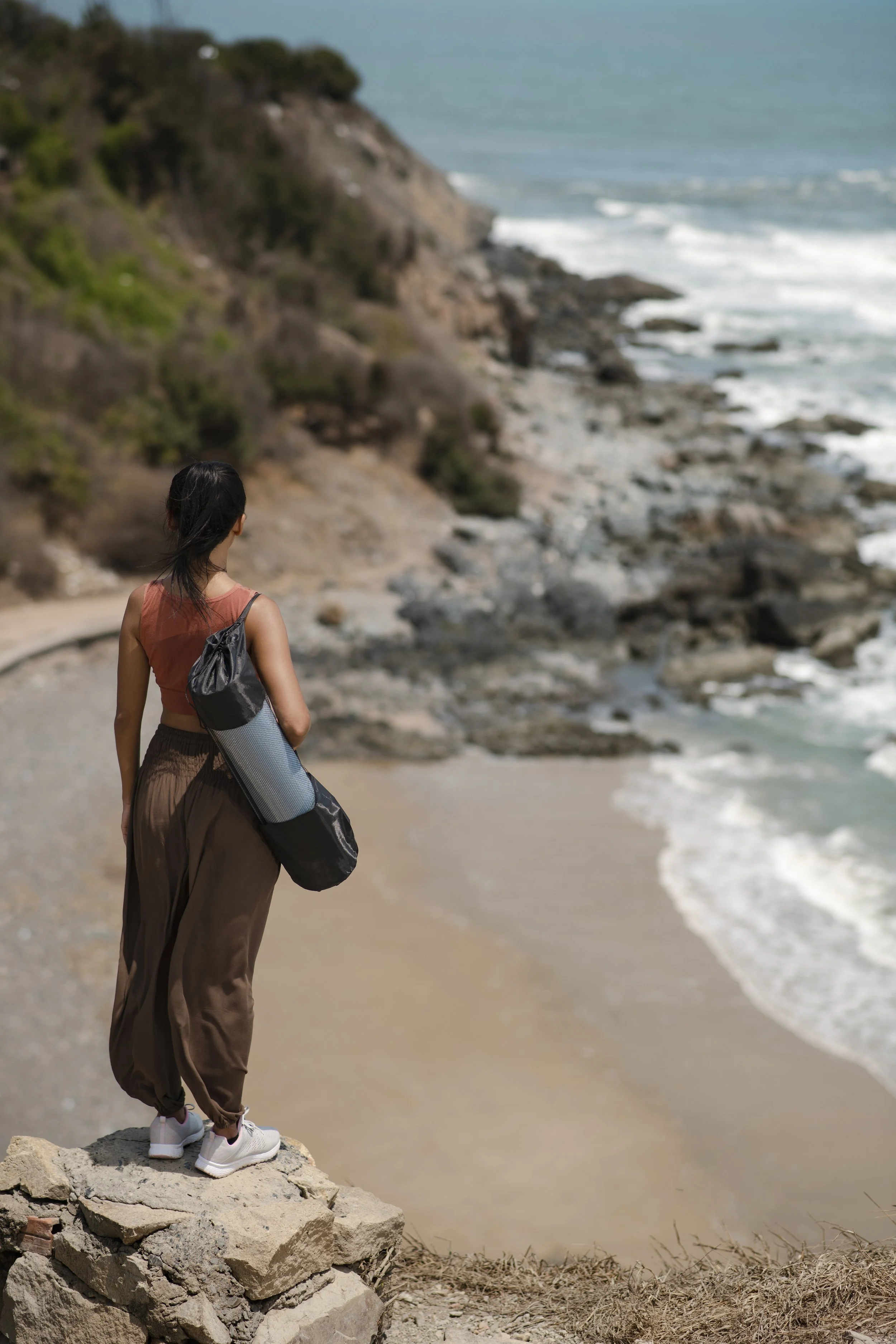 A woman standing on rocks by the beach, gazing at the ocean with cliffs in the background.