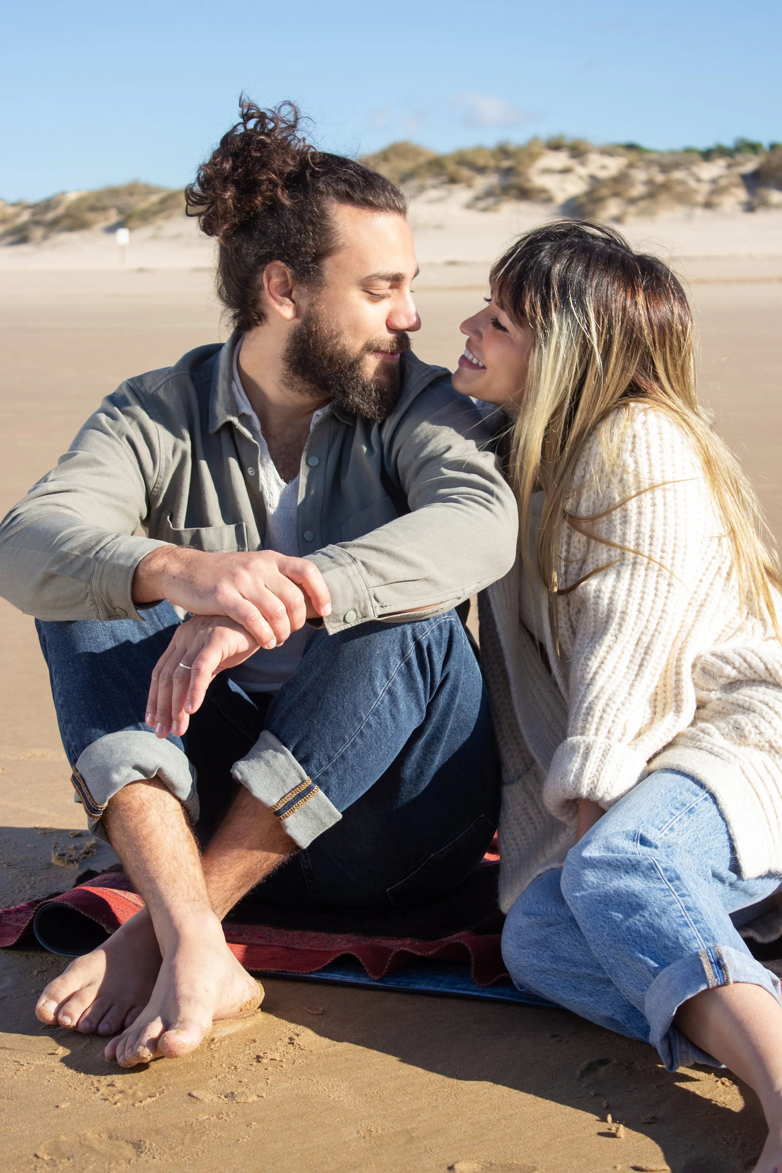 A couple sitting on a beach, smiling at each other, with sandy dunes and a clear blue sky in the background.