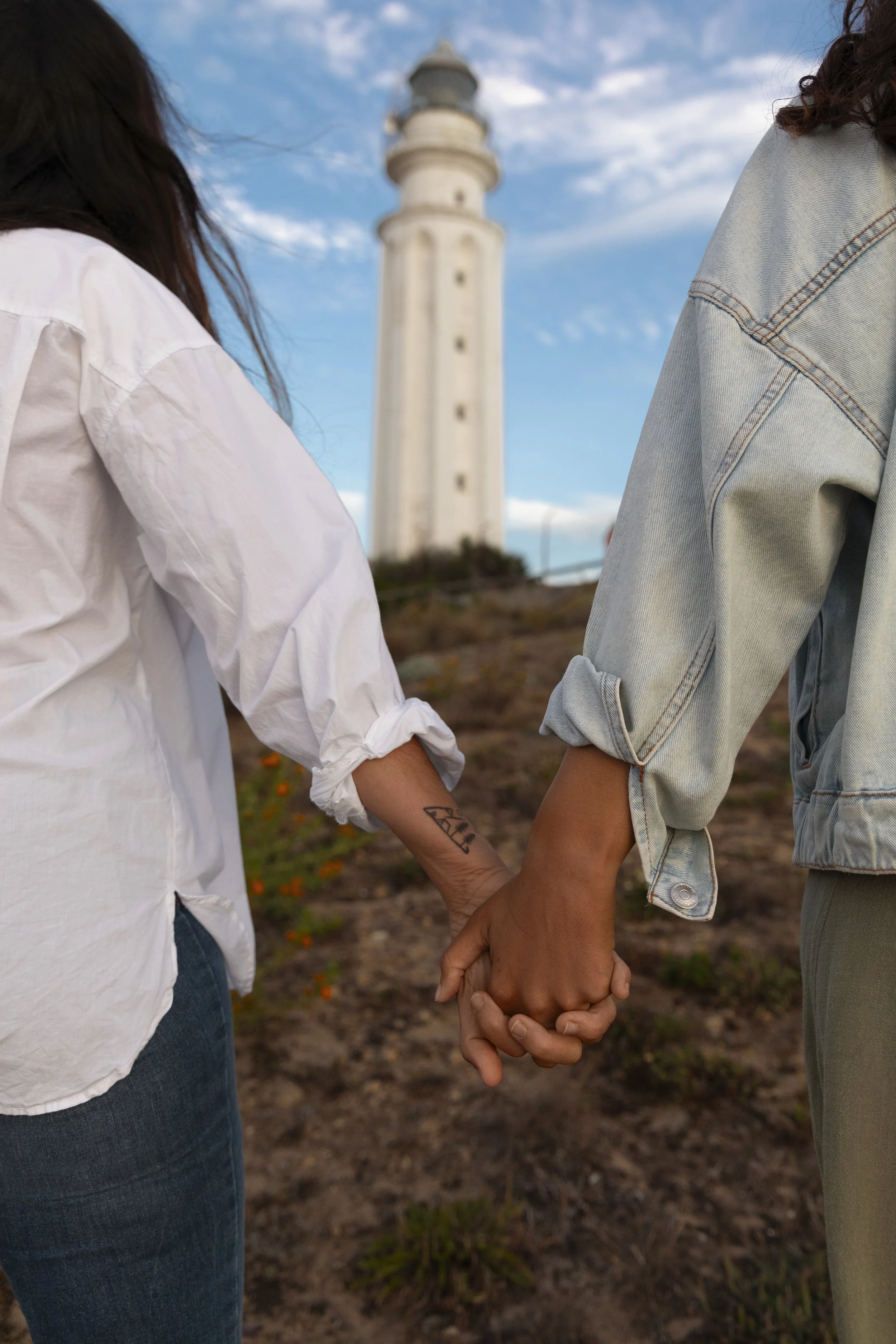 Two people holding hands outdoors with a lighthouse in the background, one wearing a white shirt and jeans, the other in a denim jacket and gray pants, during daytime with a partly cloudy sky.
