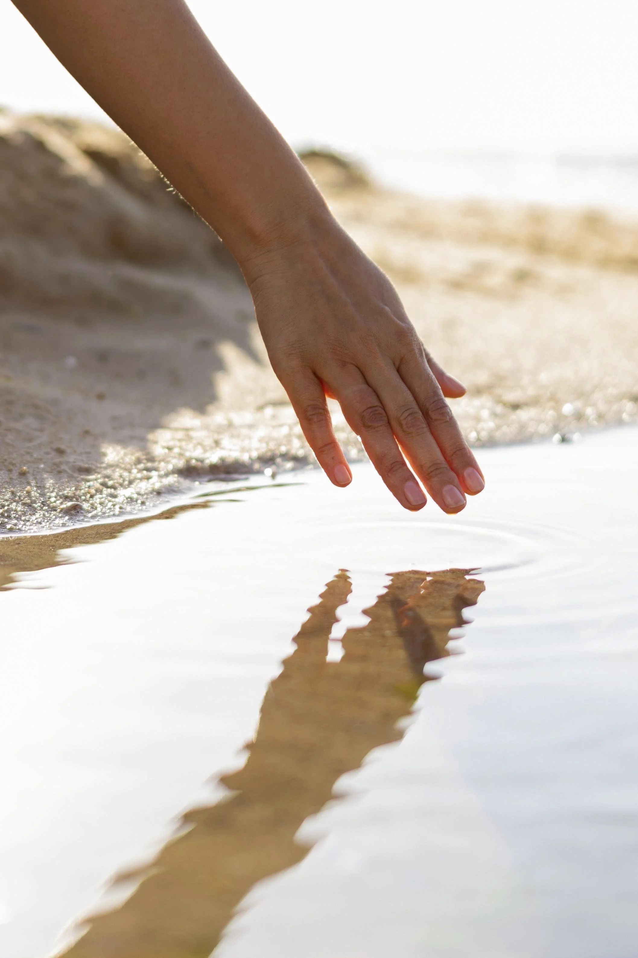 Close-up of a hand with fingers touching water at the edge of a sandy beach, with a blurred ocean background.