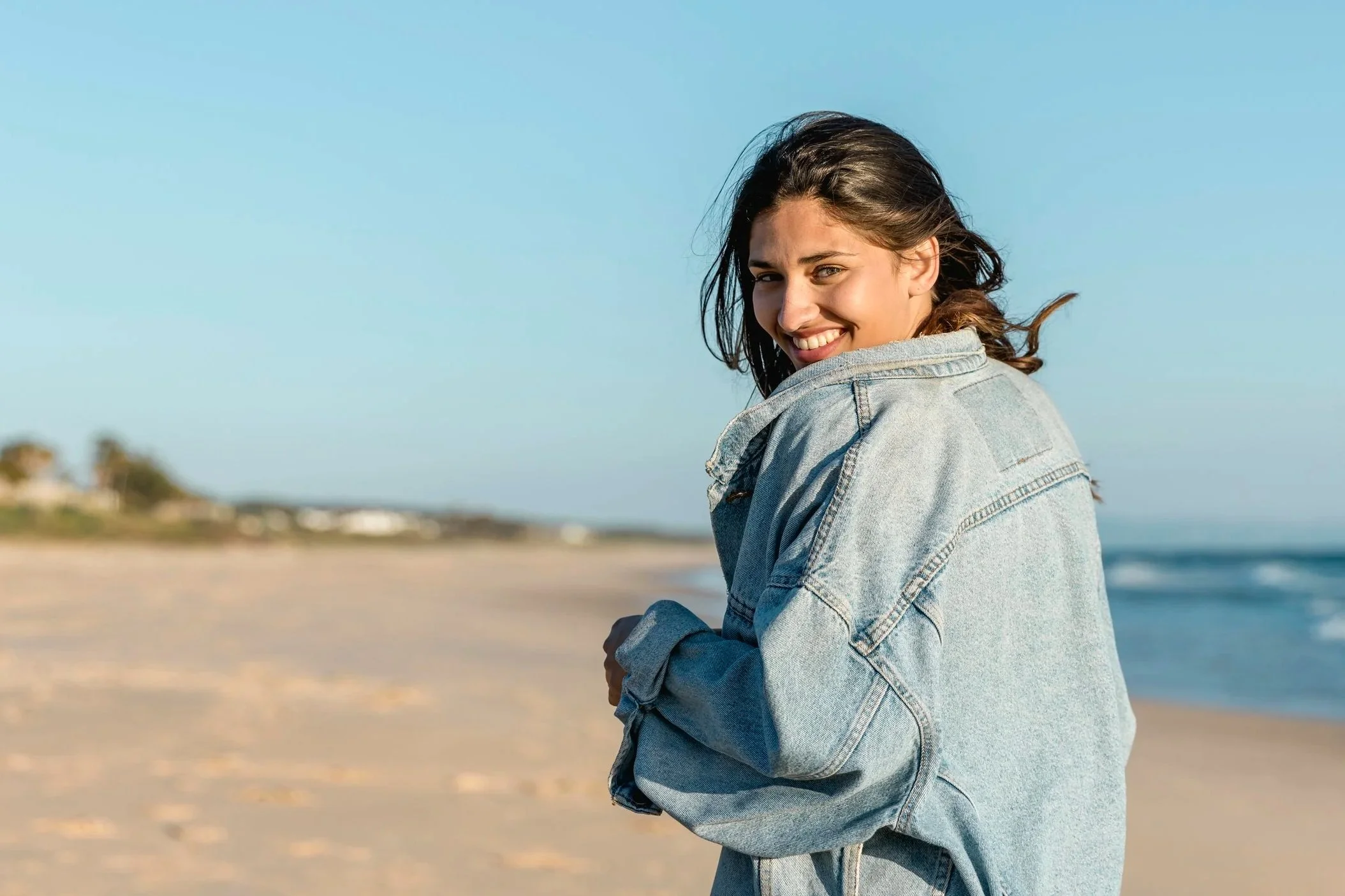 A young woman with long brown hair smiling and wearing a denim jacket at the beach with a clear blue sky.