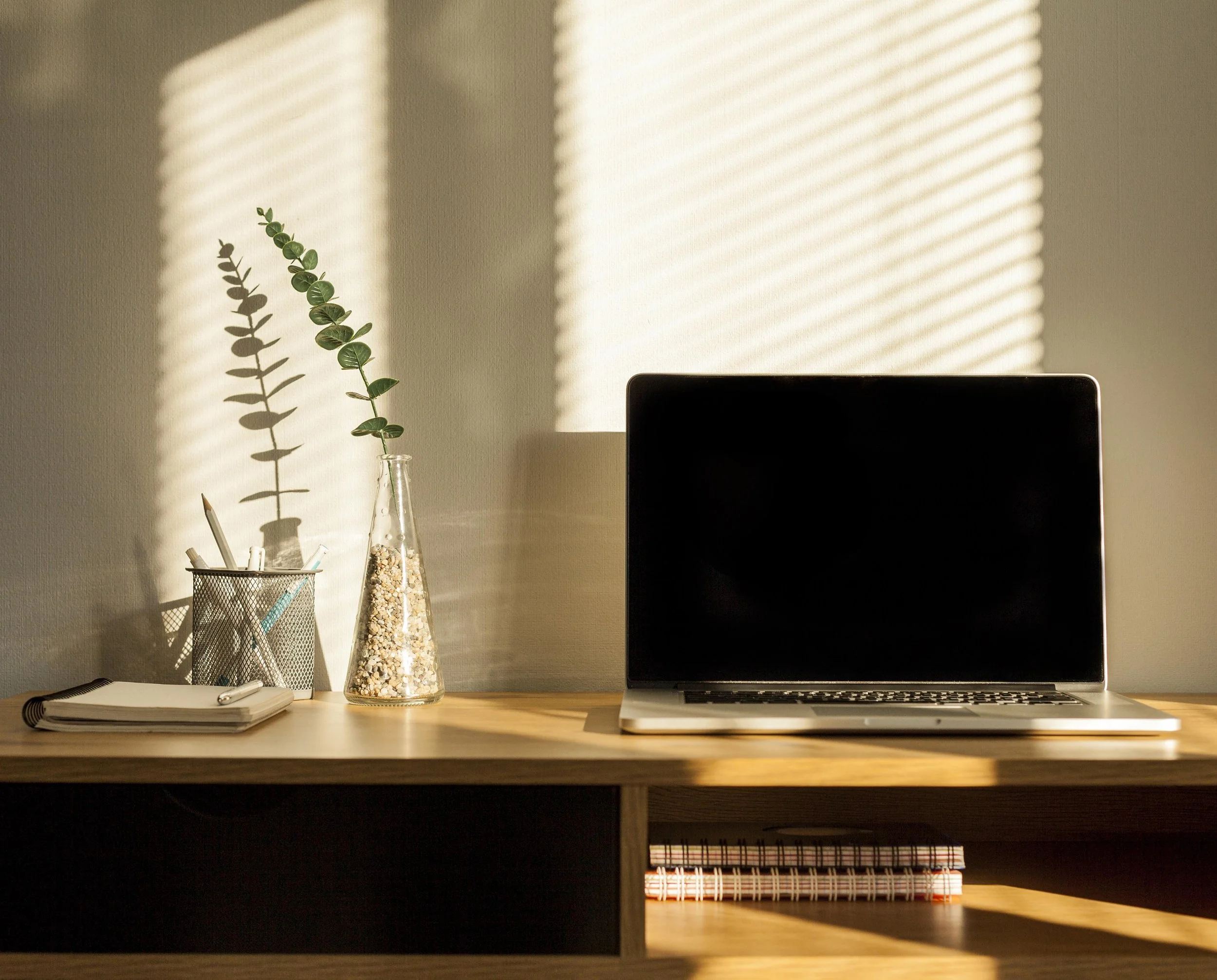 A workspace with a wooden desk, a black laptop, a spiral notebook, a pen holder with pens, a glass vase with a green plant, sunlight casting striped shadows on the wall.
