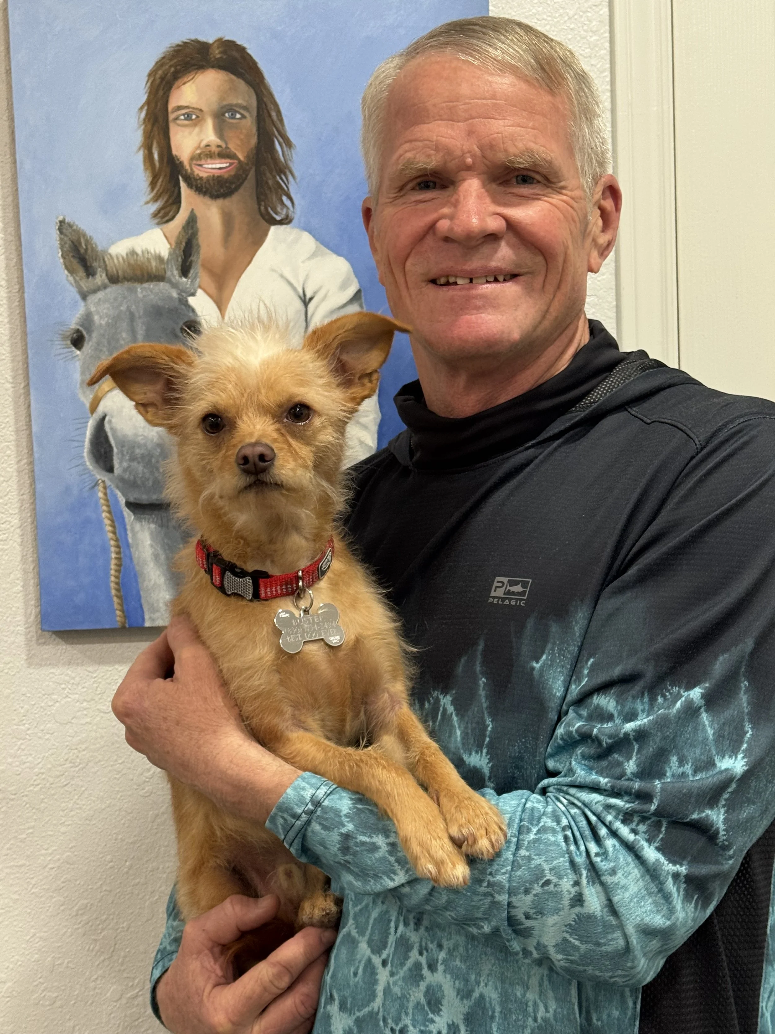 An older man with short gray hair, smiling, holding a small tan dog with pointy ears and a red collar. In the background, there is a painting of a man with long brown hair, beard, and white robe, standing next to a white horse.