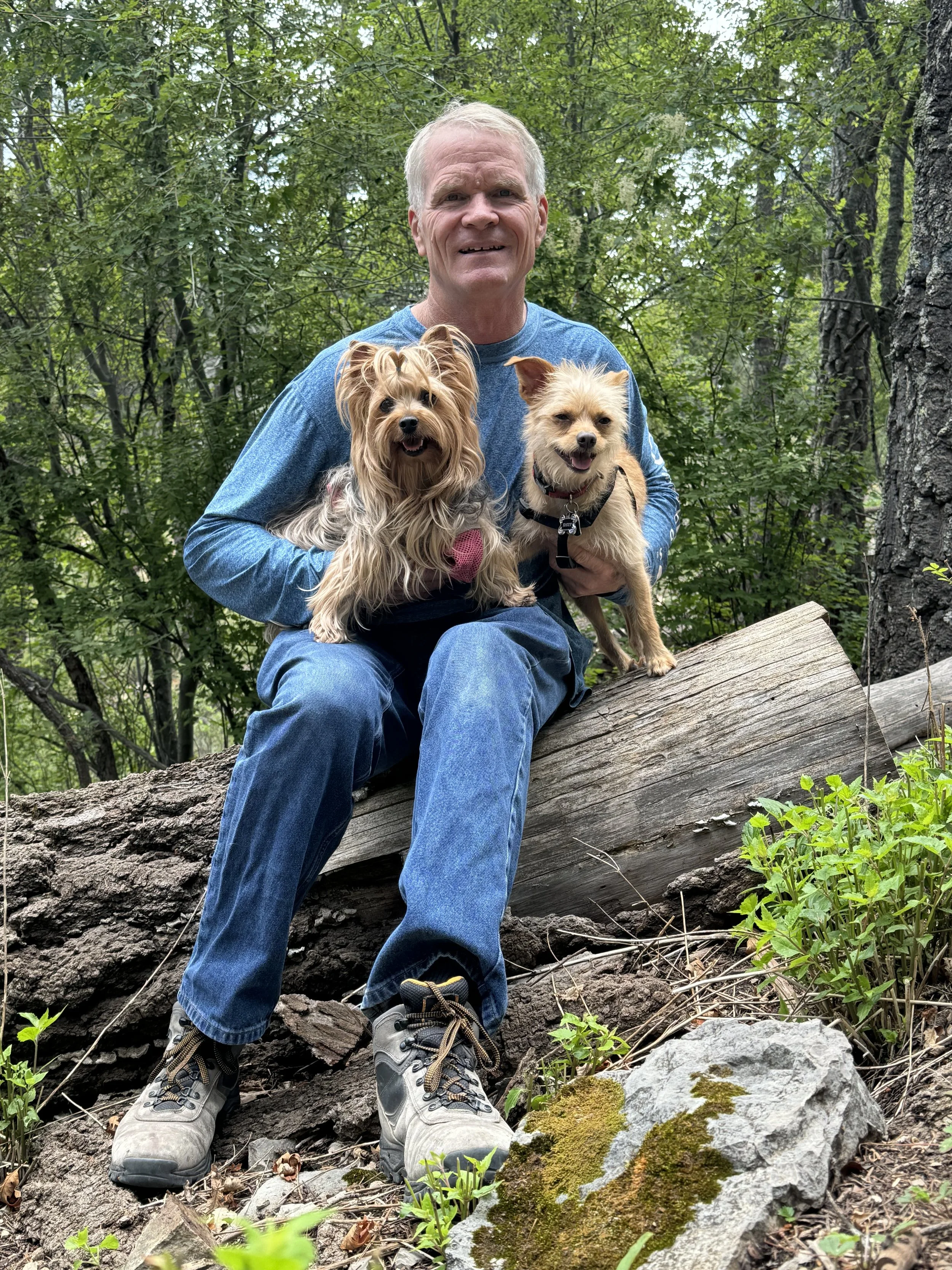 A man sitting on a fallen tree trunk in a forest, holding two dogs, smiling at the camera.