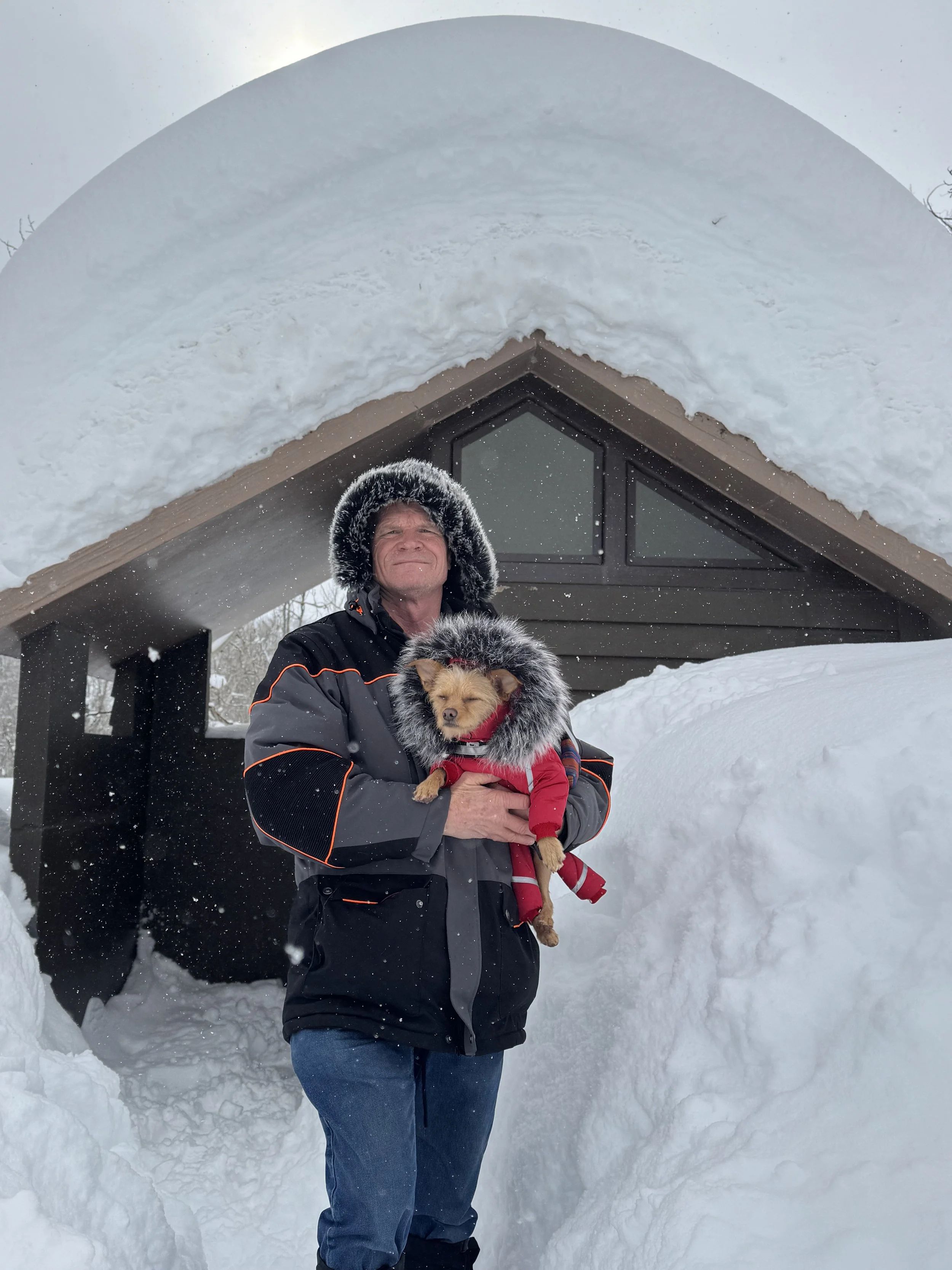 A man standing outdoors in snowy weather holding a small dog dressed in a red snowsuit. They are standing in front of a house with an arched snow-covered roof.