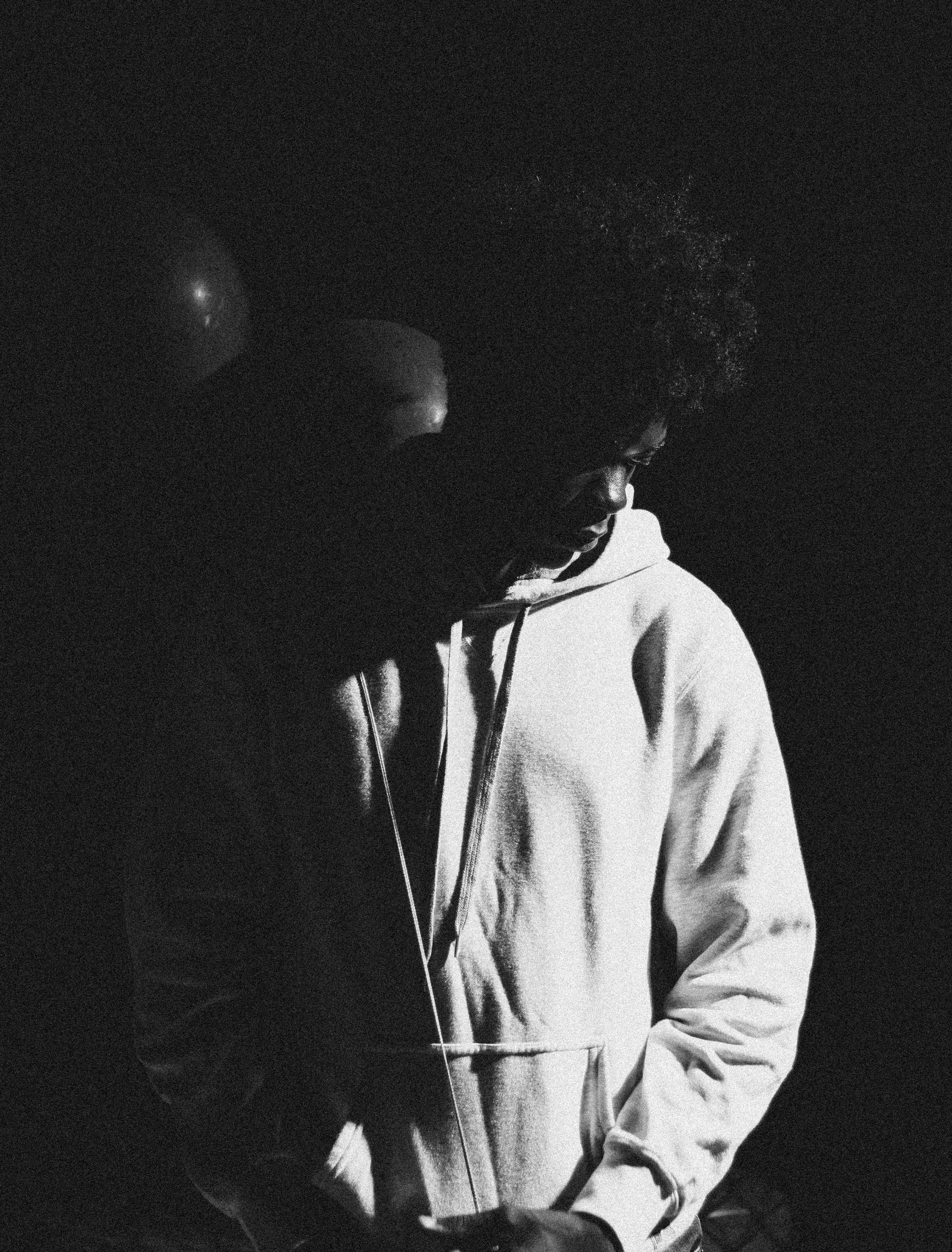 Black and white photo of a person with curly hair wearing a hoodie, standing in the dark with head tilted downward.