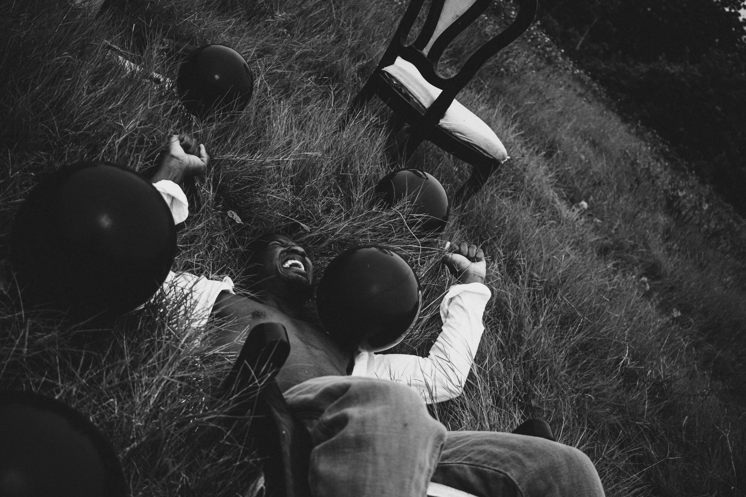A black man lying on the grass, smiling, surrounded by four black balloons, with a wooden chair nearby in an outdoor setting.