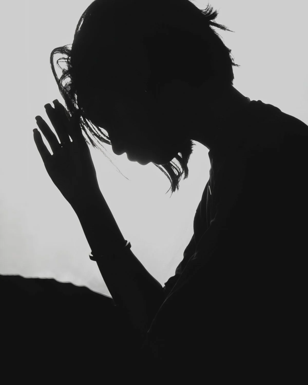 Silhouette of a person with head bowed and hand raised near temple, against a bright background.