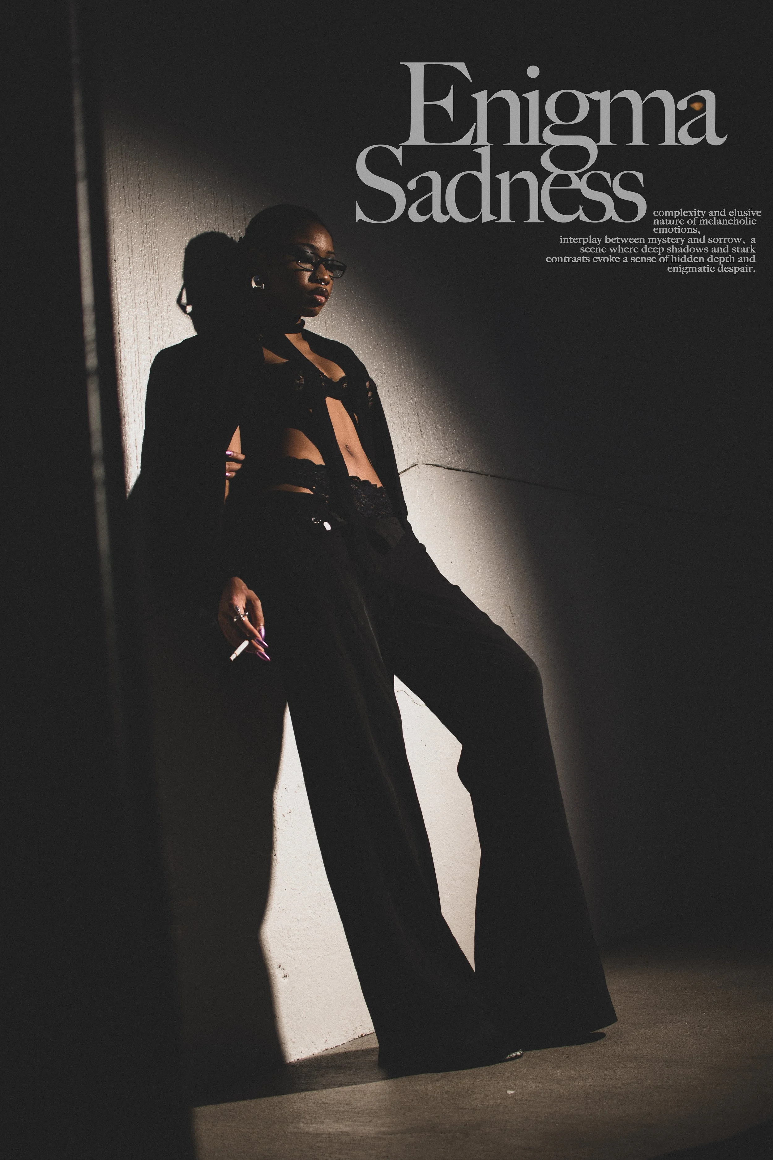 A woman standing against a white wall in a dark room, wearing black clothing and glasses, holding a cigarette.