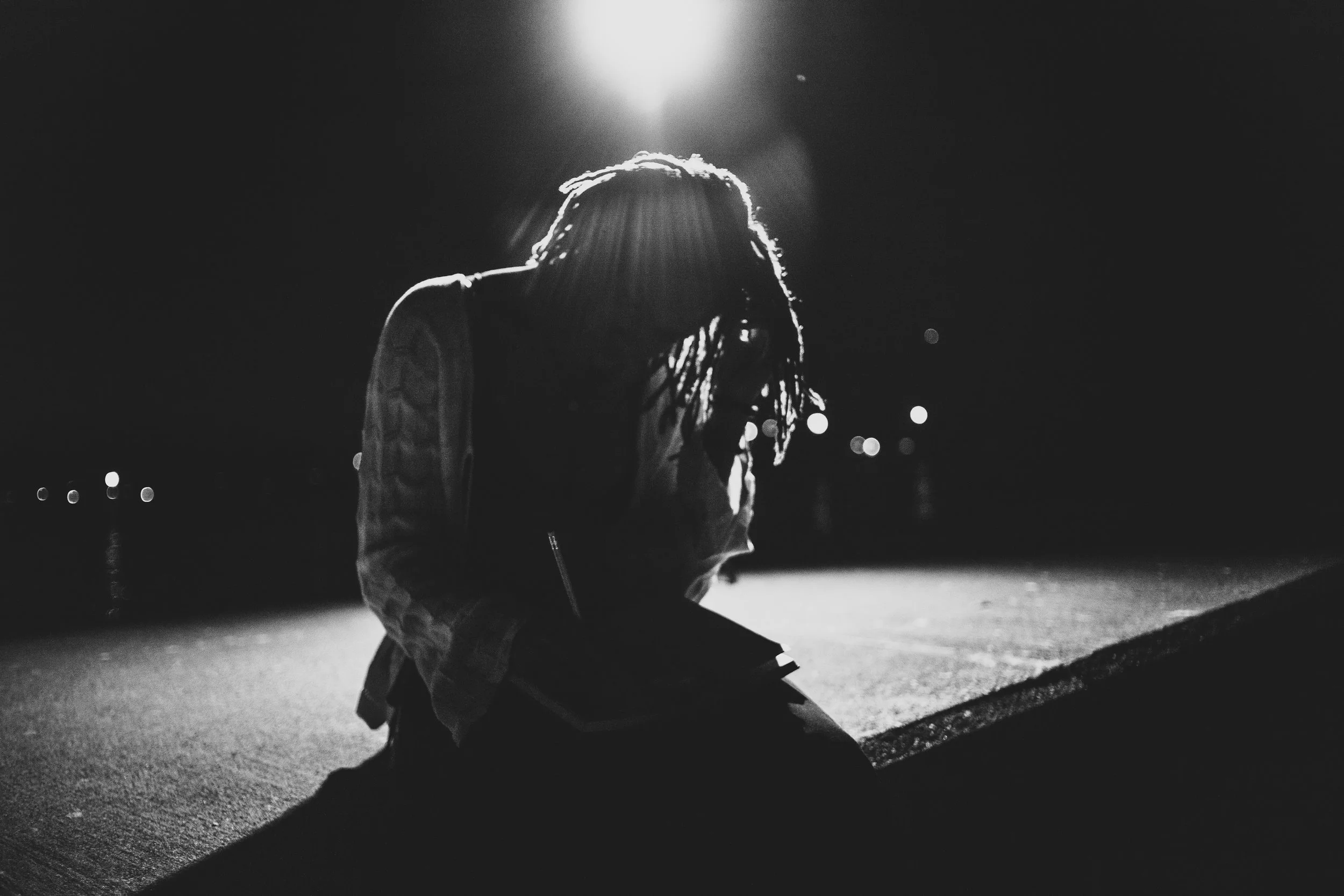 A person with dreadlocks wearing glasses, sitting on the ground at night, illuminated by a bright streetlight behind them.
