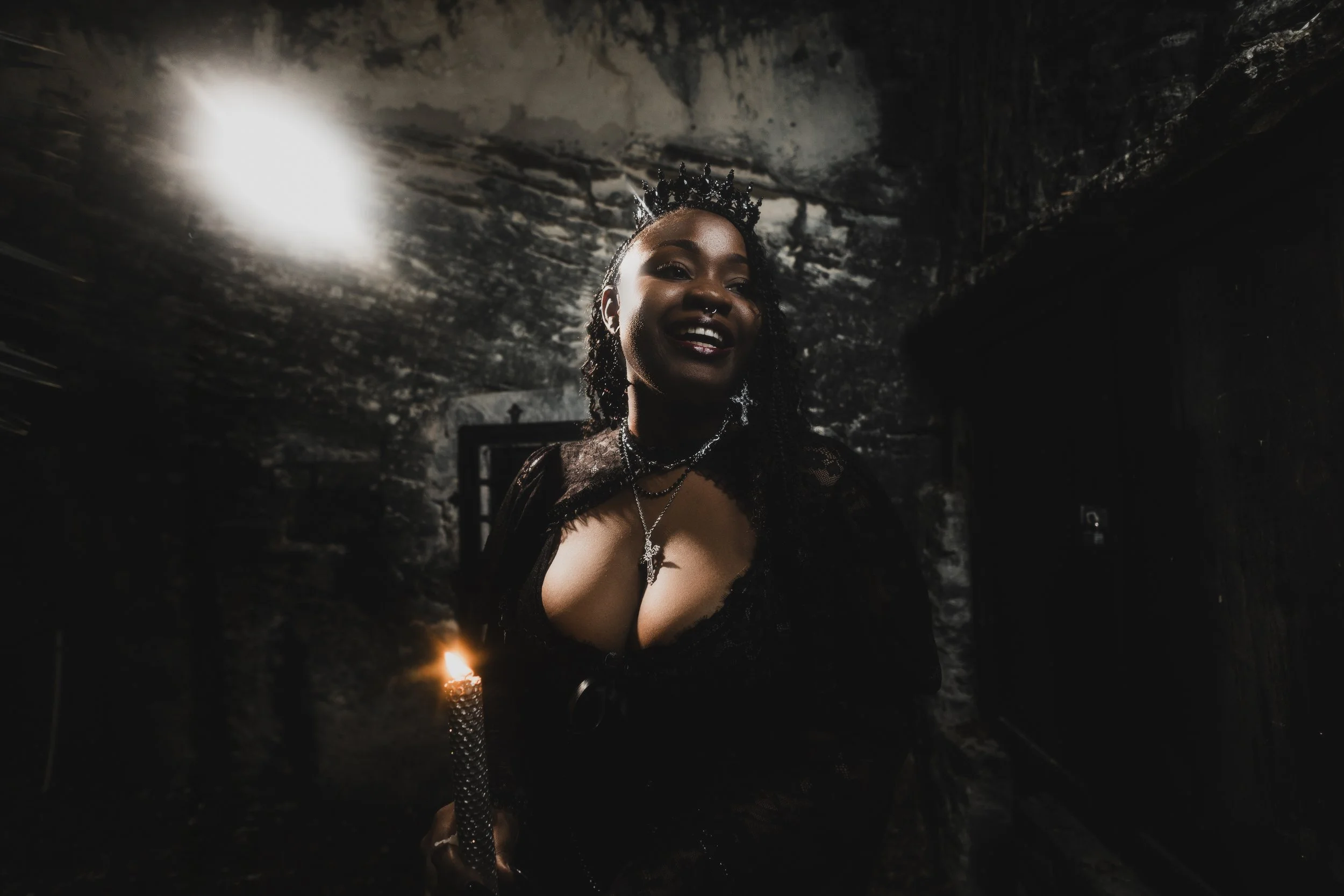 A woman in dark clothing with a crown and jewelry holds a lit candle in a dimly lit, rocky room, smiling.