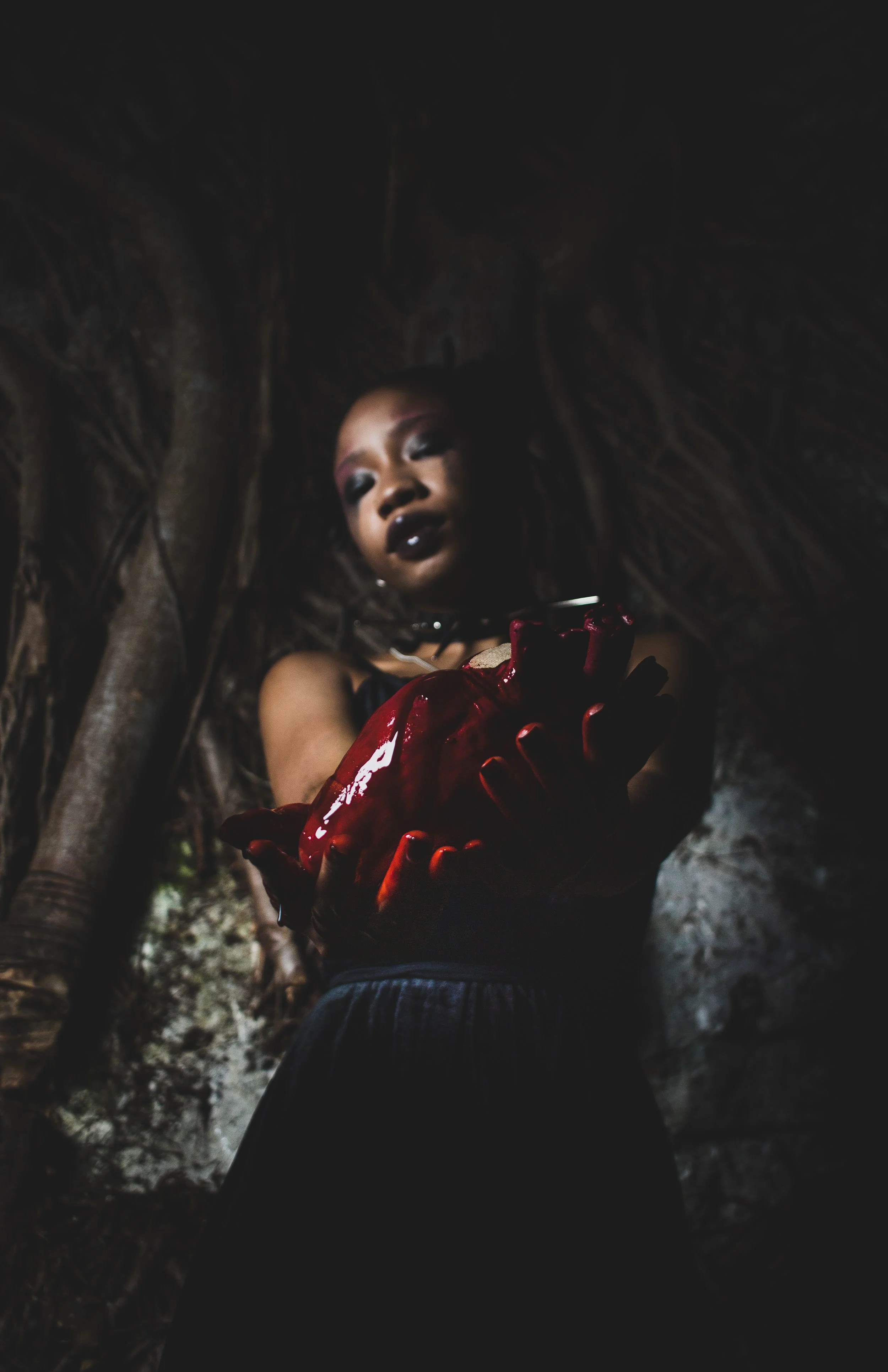 A woman with dark makeup and gloves is holding a large, red, possibly artificial or artistic, anatomical heart in a dark, textured environment.
