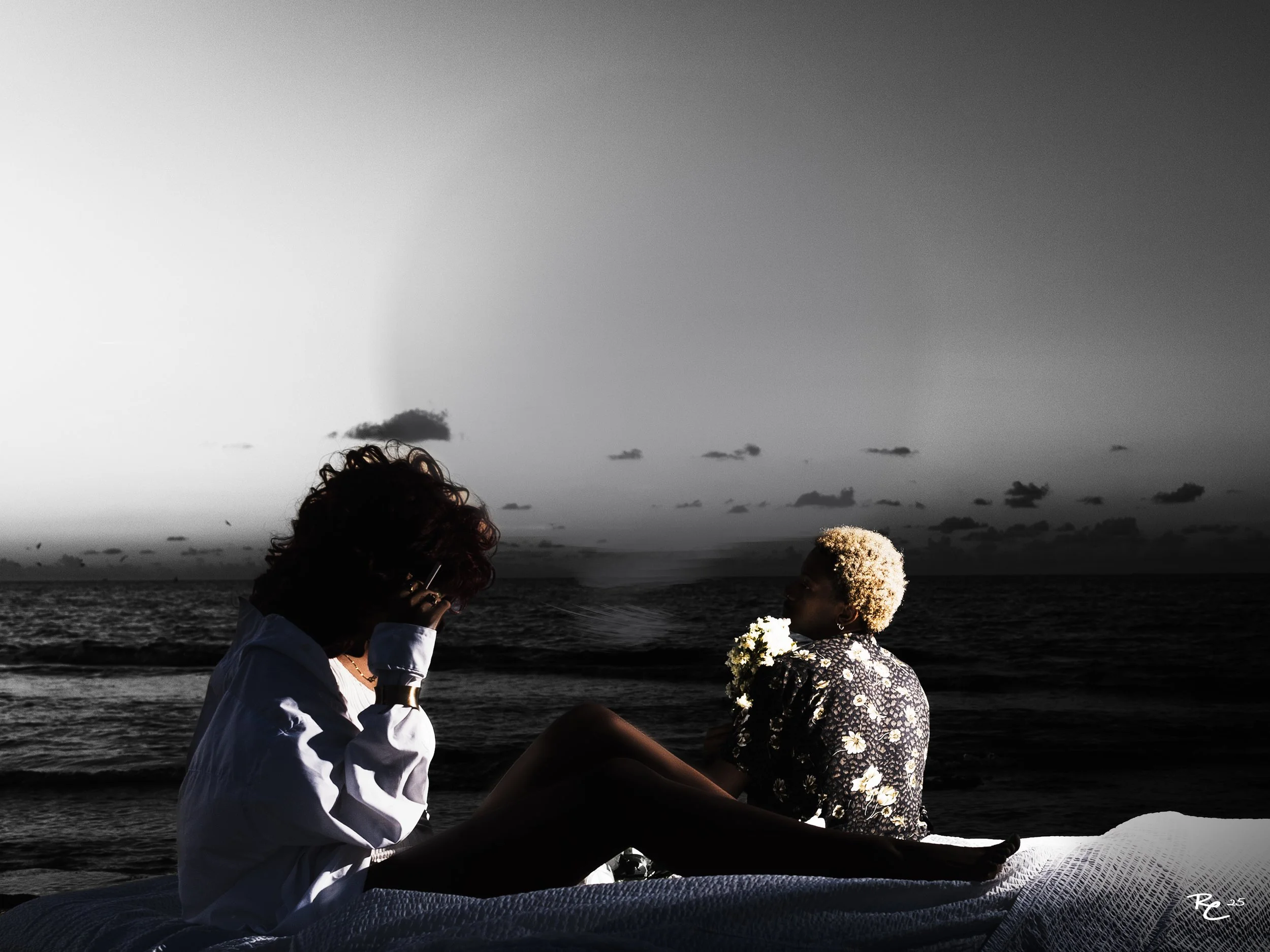 Two women sitting on a blanket on the beach during sunset. One woman with curly hair is talking on a phone, wearing a white shirt. The other with short, blond hair is wearing a floral top, holding a bouquet of flowers, looking out at the ocean.