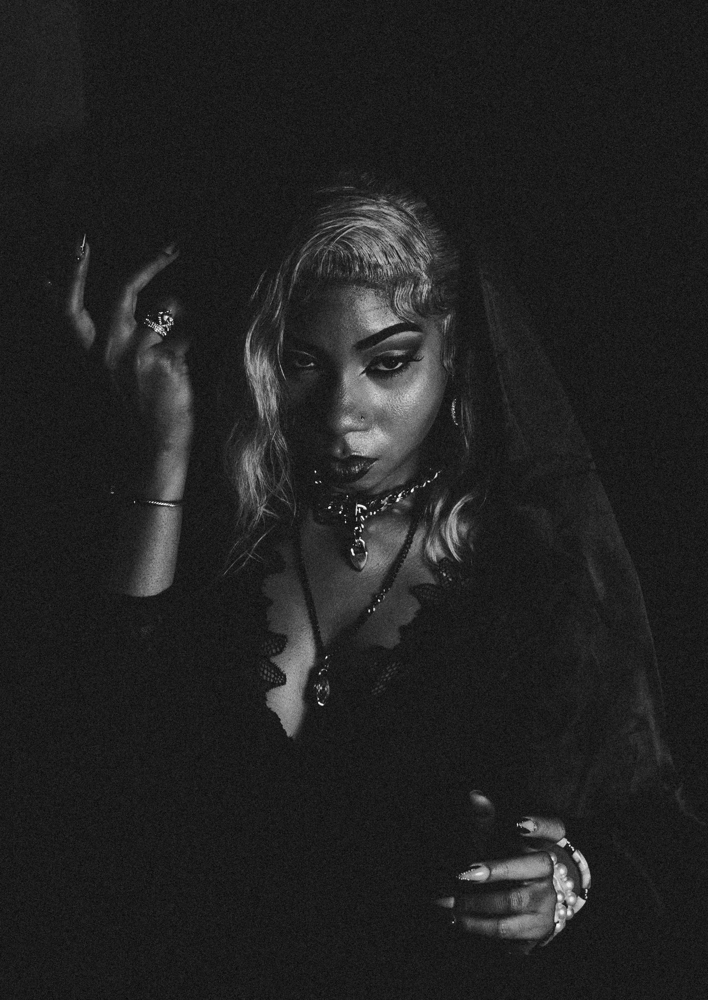 A woman with styled wavy hair and dark makeup, wearing jewelry, in a dark setting with dramatic lighting.