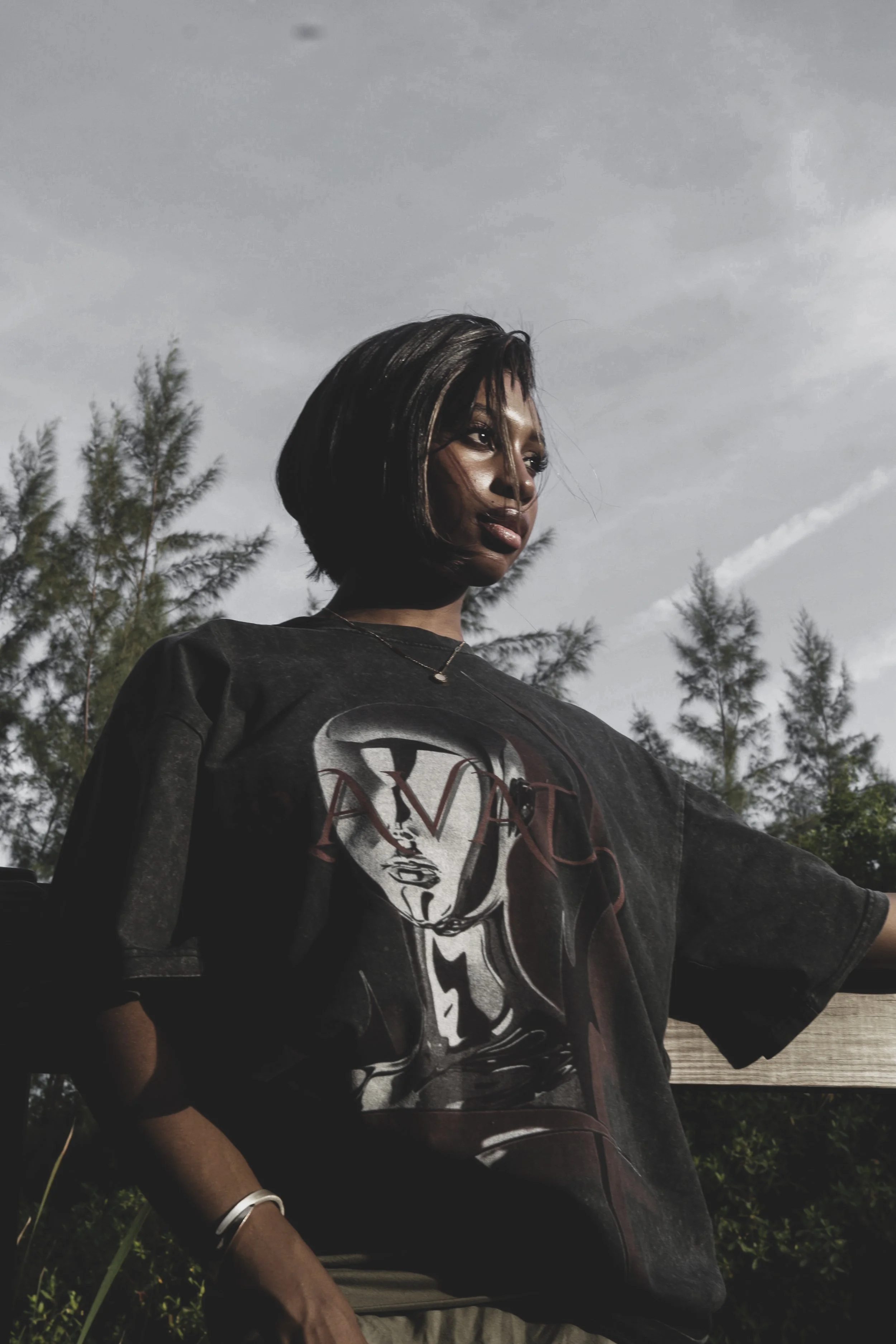 A woman with short black hair and dark lipstick looks thoughtfully into the distance outdoors, wearing a black graphic T-shirt with a woman's face and the word 'LOVE' on it, trees and a cloudy sky in the background.
