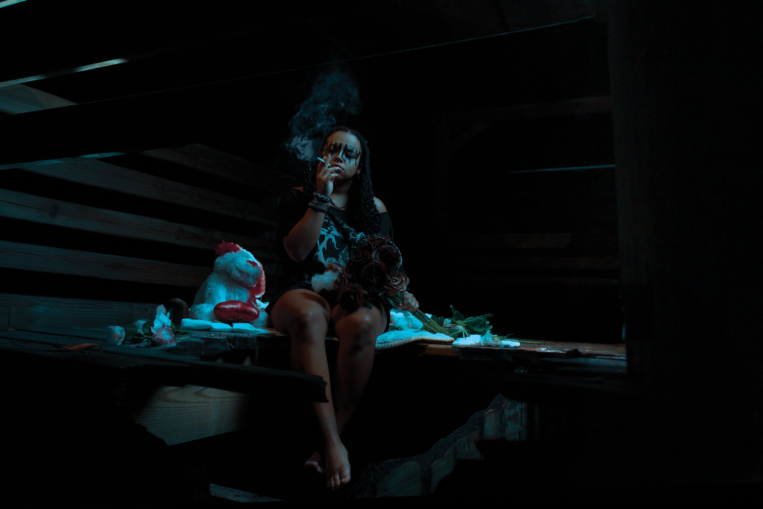 A woman sitting on a wooden platform in a dark space, smoking a cigarette with smoke rising from it. She is surrounded by a stuffed animal, roses, and other objects. The scene has a moody, nighttime atmosphere with low lighting.