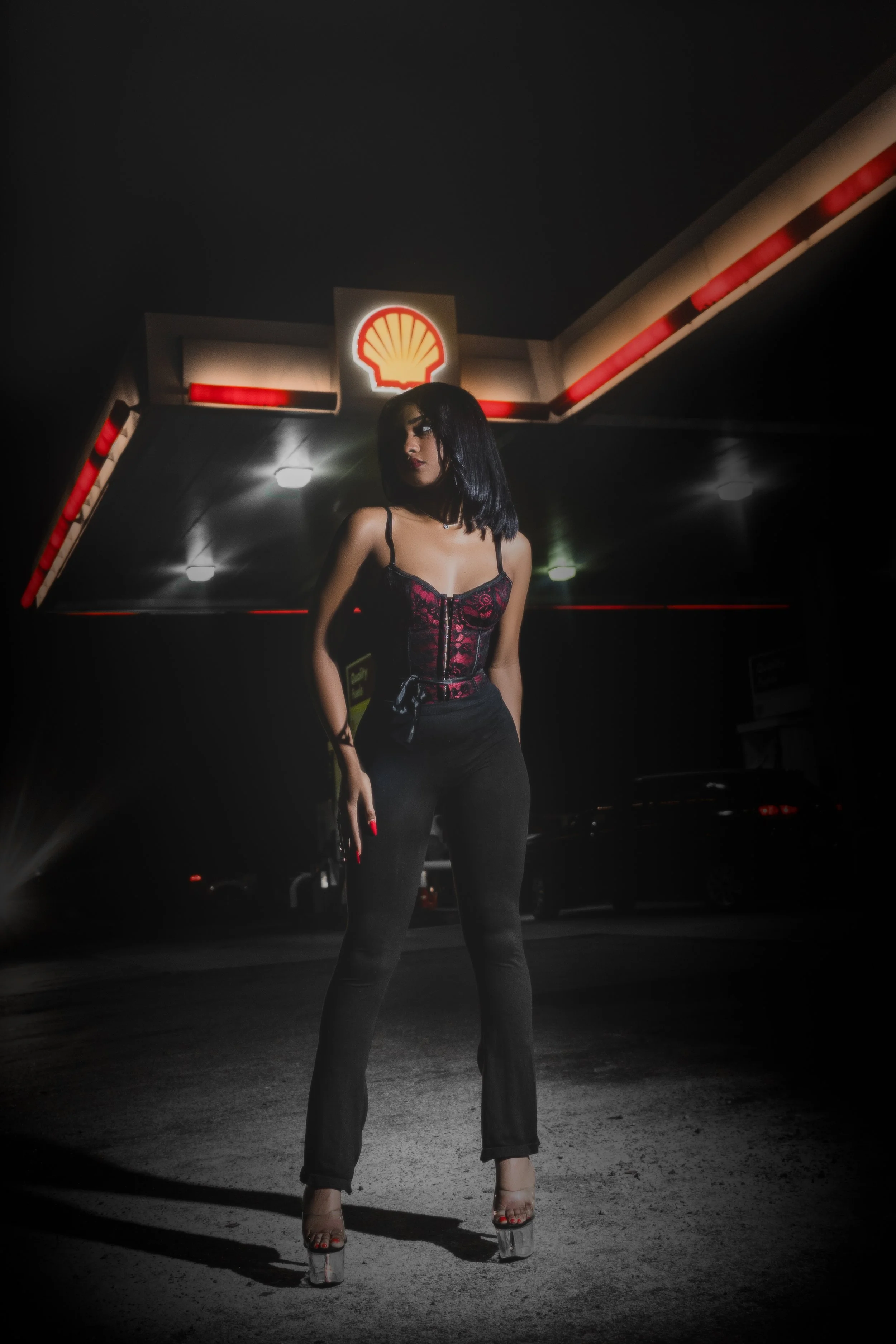 A woman with shoulder-length black hair and red lipstick stands outside a Shell gas station at night, wearing a black and red lace top, black pants, and clear high heels.