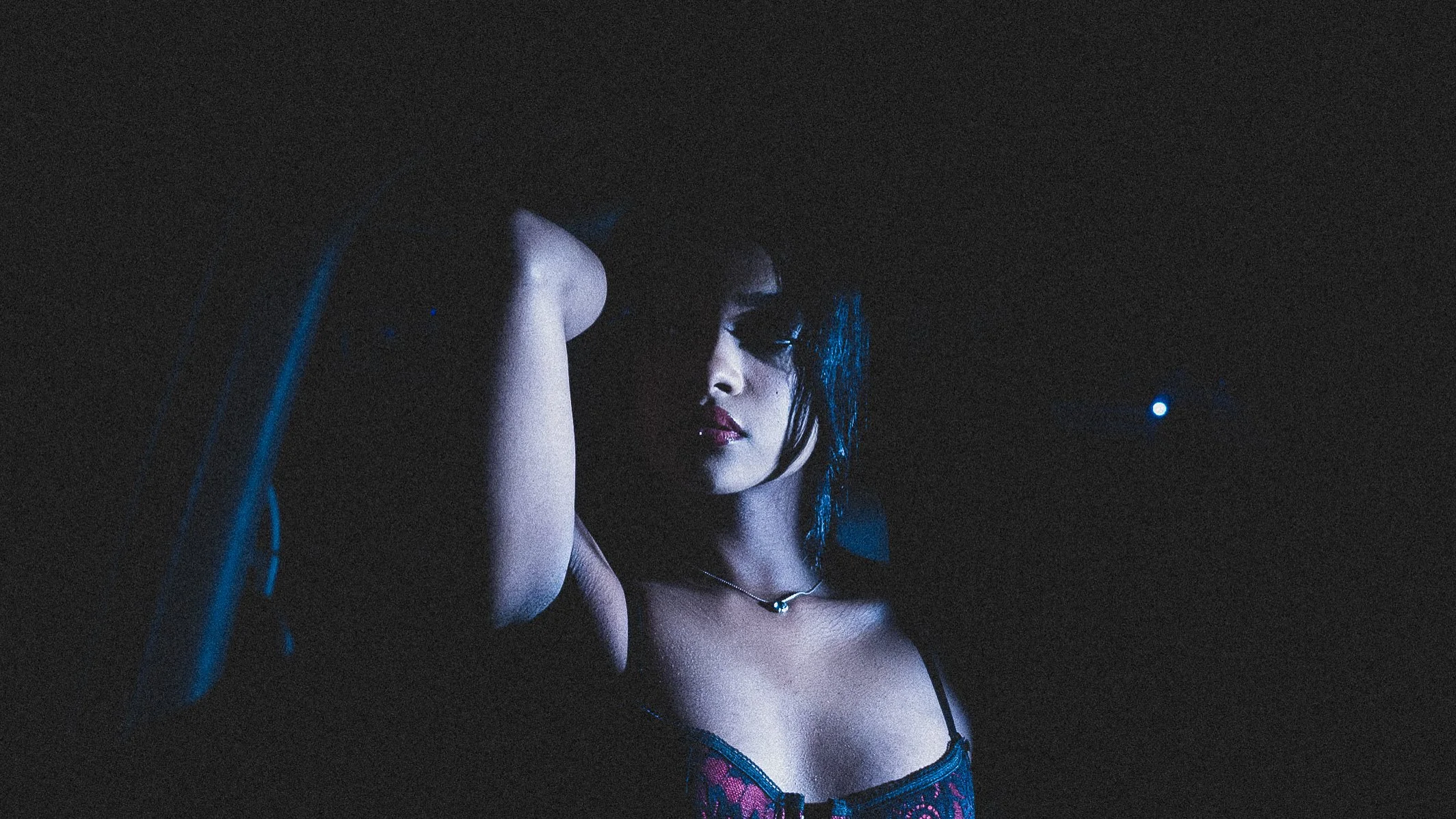 A woman with dark hair and makeup, wearing a black and red lace dress, is sitting in a dimly lit environment with her arm resting on her head, illuminated by a soft light.