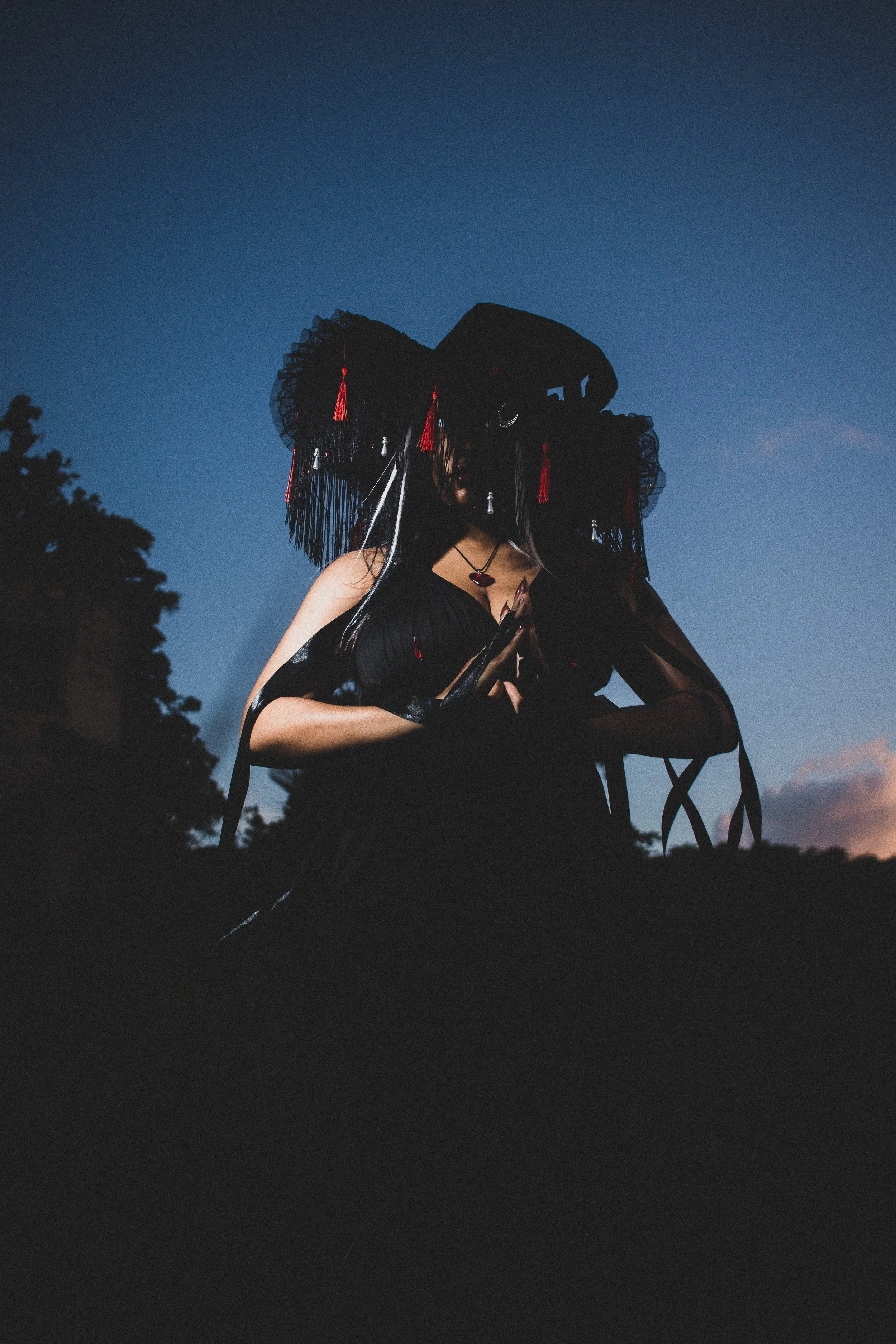A person dressed in black with a large, ornate black hat adorned with red tassels and ribbons, standing outdoors during sunset or twilight, with a darkened landscape and trees in the background. They are looking down with their hands clasped in front of them.