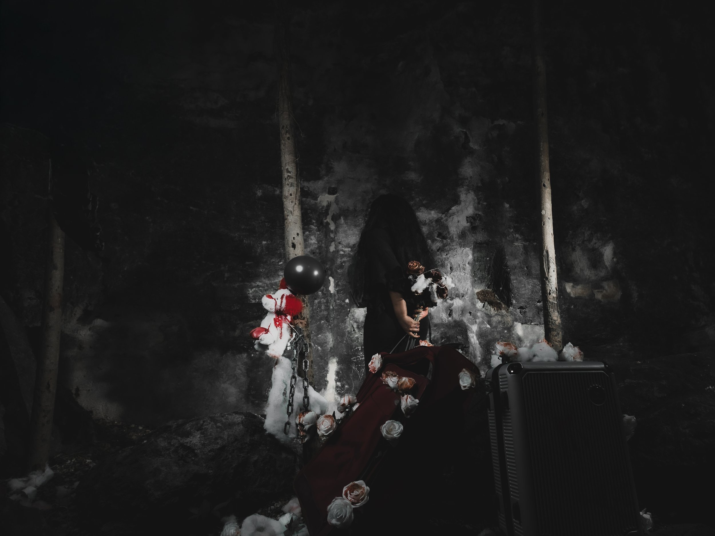 A person dressed in black with a black hood covering their face, standing in a dark, cave-like environment. They hold a bouquet of flowers, surrounded by cotton-like material, with a black balloon attached to chains on the left. There is a black case or box on the ground in front of them.