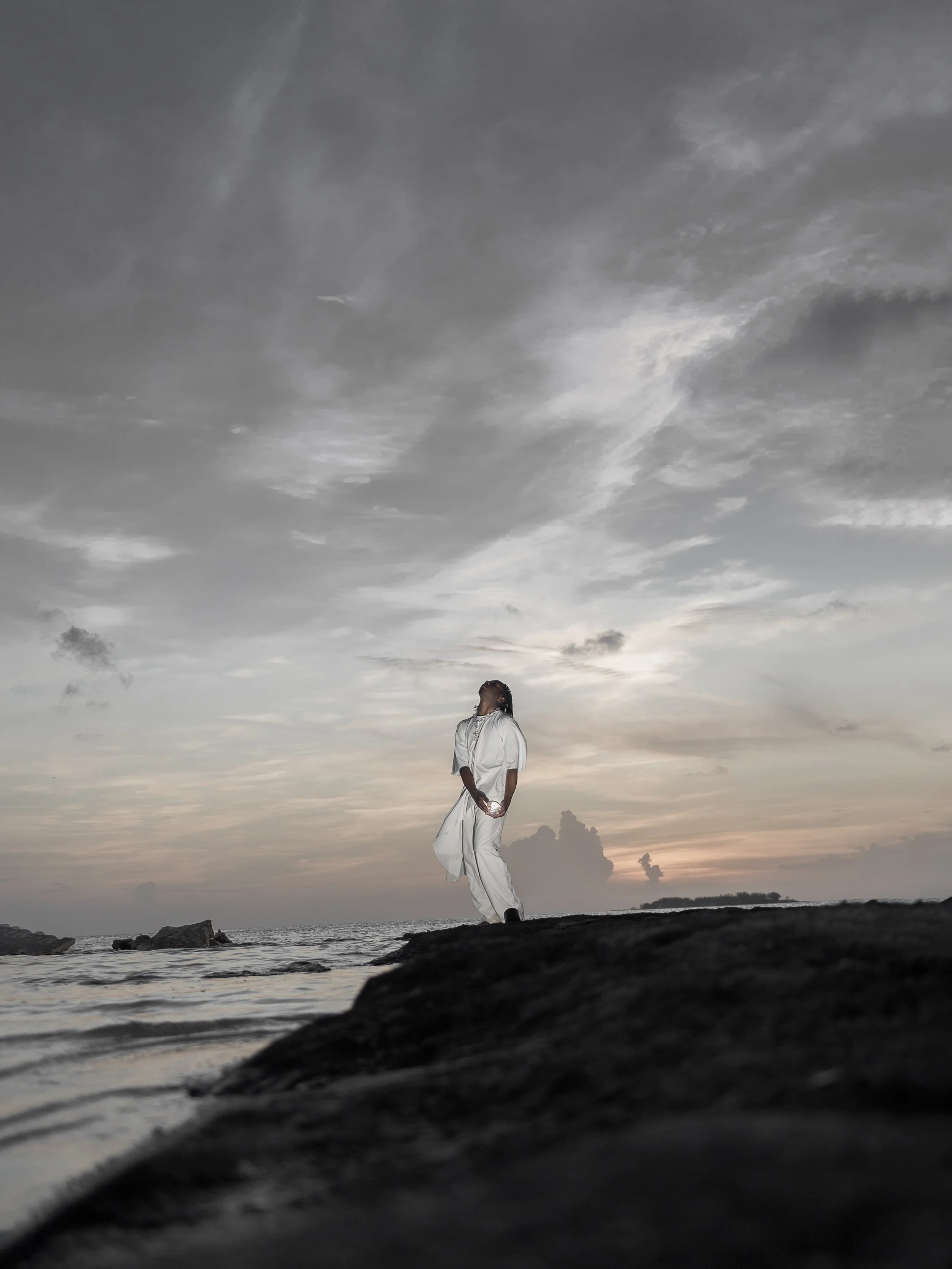 Person in white clothing standing on a rocky beach during sunset or sunrise, with a cloudy sky above and the ocean in the background.