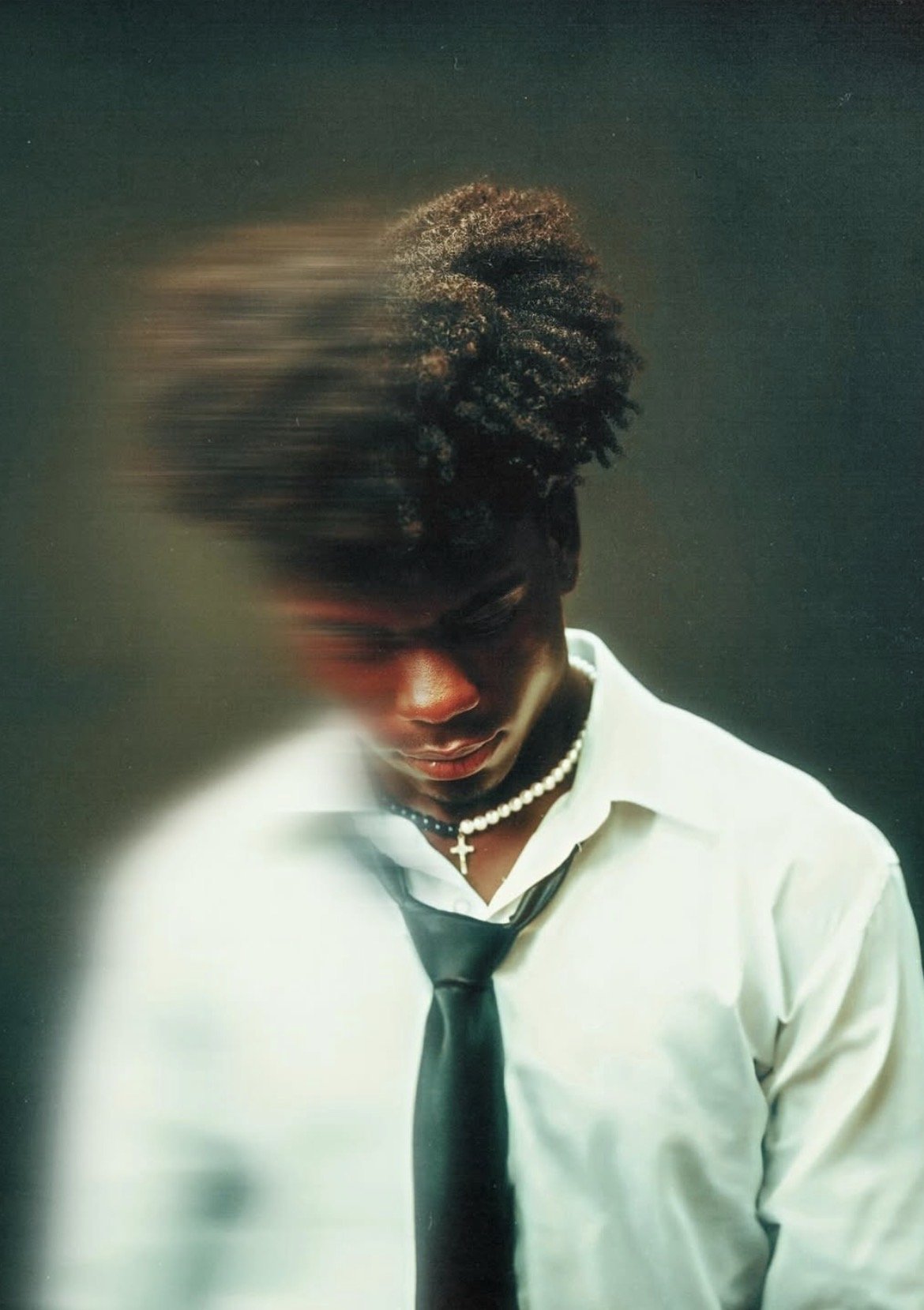 Portrait of a young man wearing a white shirt, black tie, and pearl necklace with a cross pendant, against a dark background, with a motion blur effect on the left side of his face and hair.