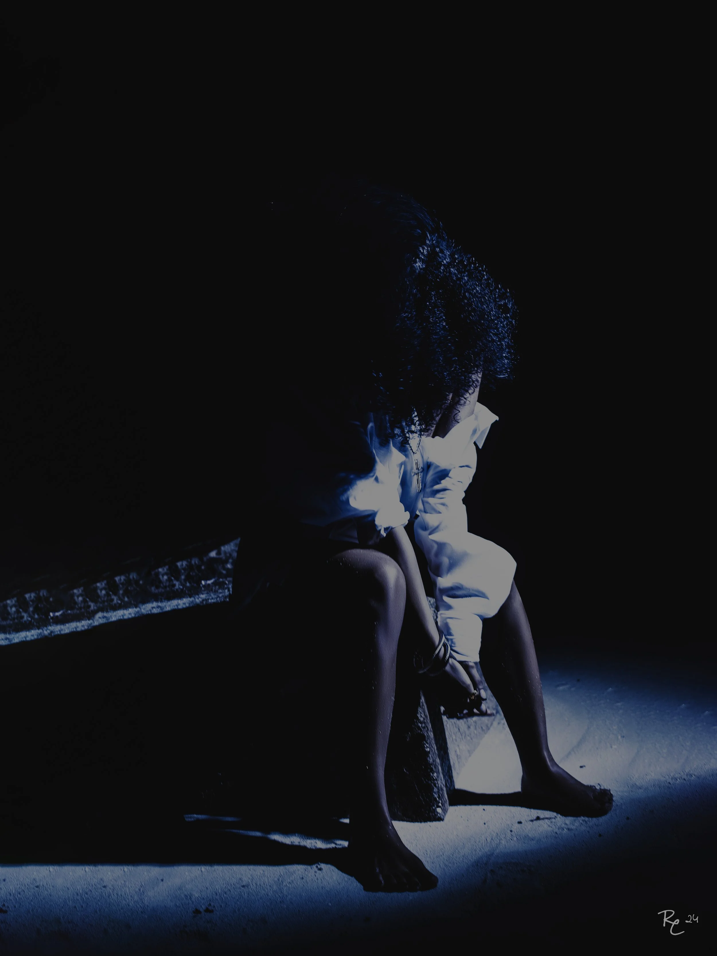 Person sitting on a bench at night, with a white jacket, illuminated by a small light, dark background.