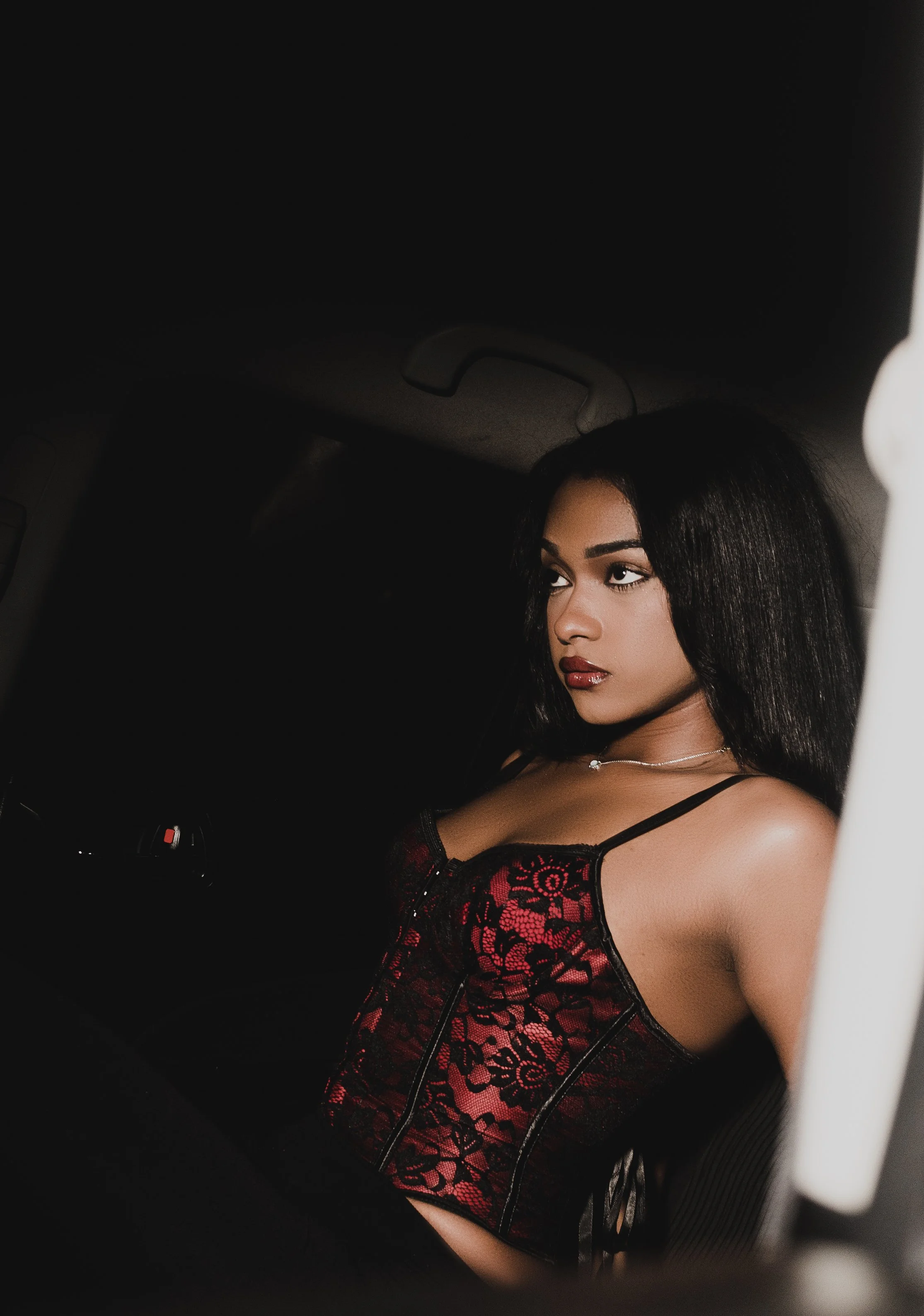 A young woman wearing a black and red lace corset top with spaghetti straps sitting in a car at night.