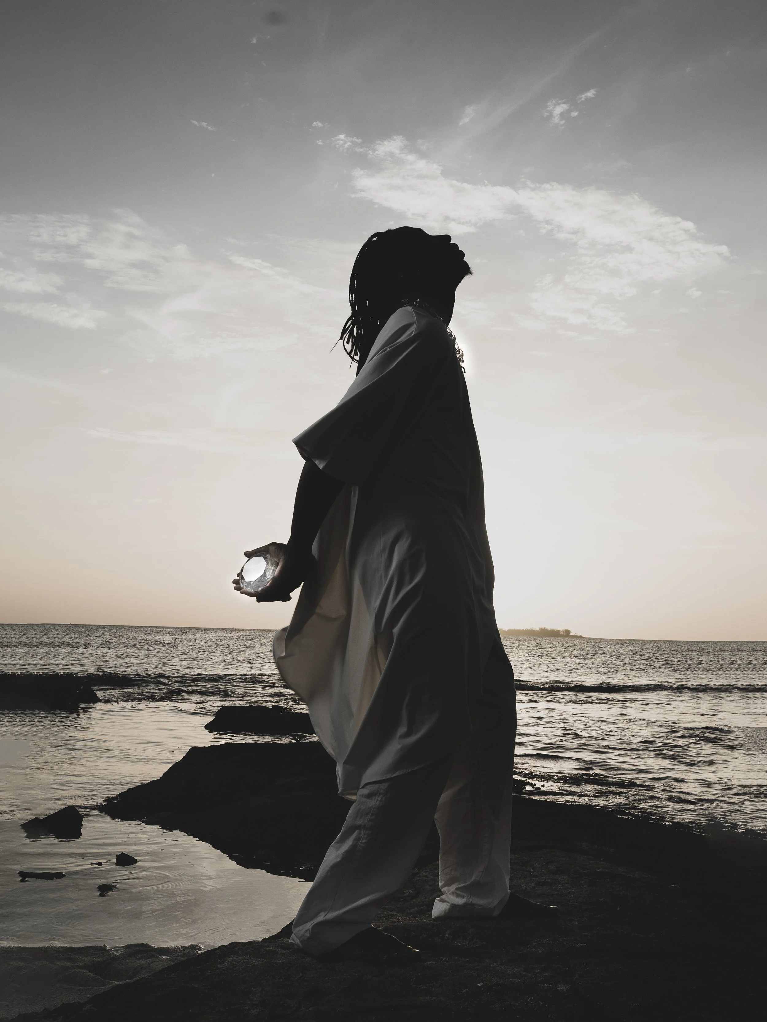 Silhouette of a woman standing on rocks by the ocean during sunset, holding a crystal ball behind her back.