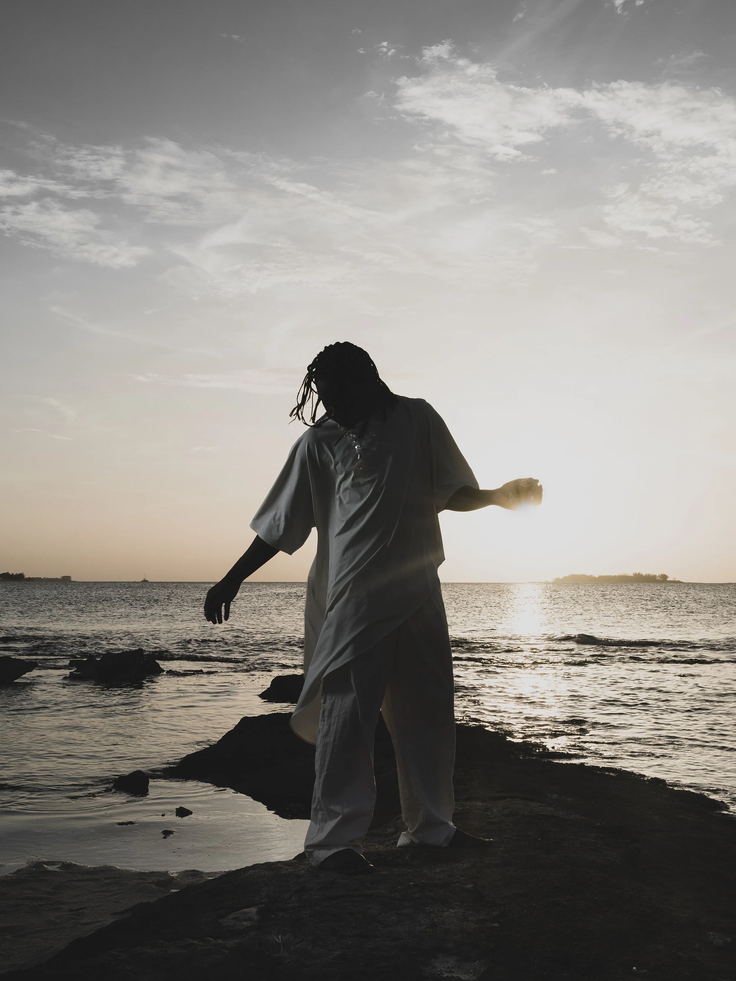 Silhouette of a person with dreadlocks standing on rocks near the water at sunset.