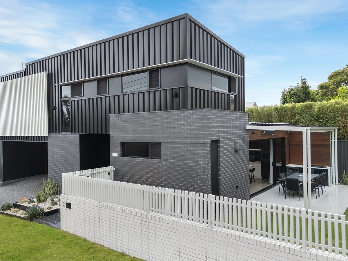 Modern multi-story house with black, gray, and white exterior, featuring a patio with outdoor dining area and a garden with desert plants, enclosed by a white fence.