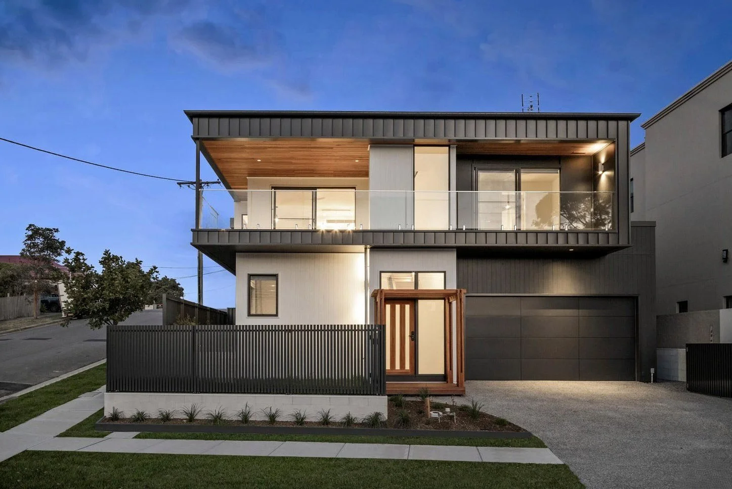 Modern two-story house with a black garage door, glass balcony, and a wooden front door, illuminated in the evening with a clear blue sky above.